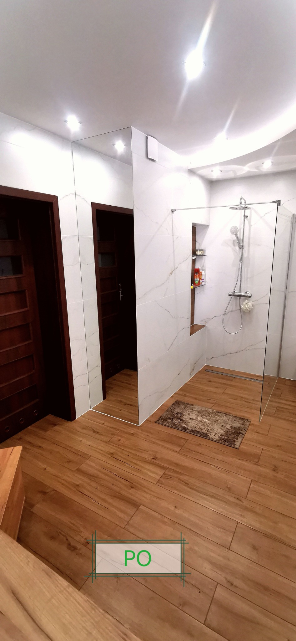 Nowoczesna łazienka z kabiną prysznicową typu walk-in, wykończona białymi płytkami imitującymi marmur i drewnopodobną podłogą, z ukrytą półką w ścianie i dużym lustrem.