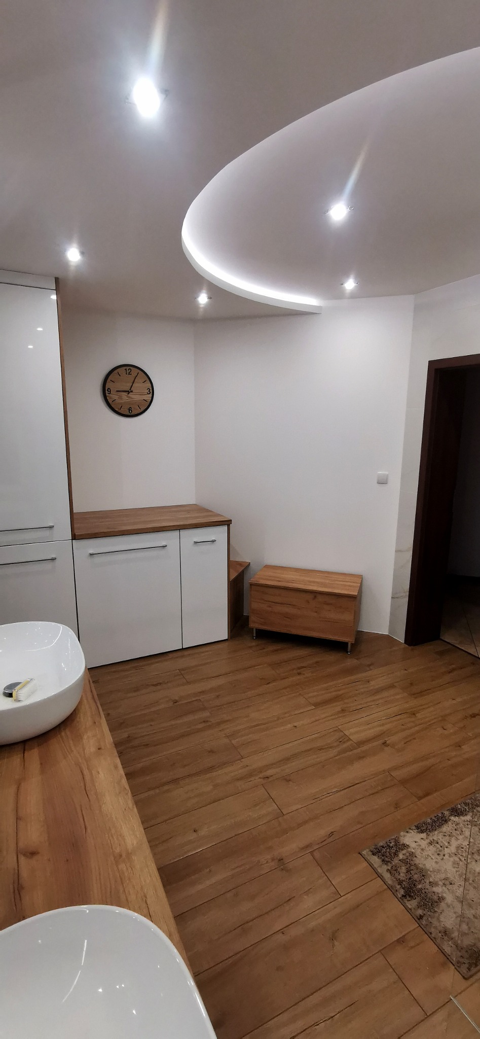 Nowoczesna łazienka z drewnianą podłogą imitującą panele, białe szafki z drewnianym blatem, okrągły zegar na ścianie i owalna umywalka.