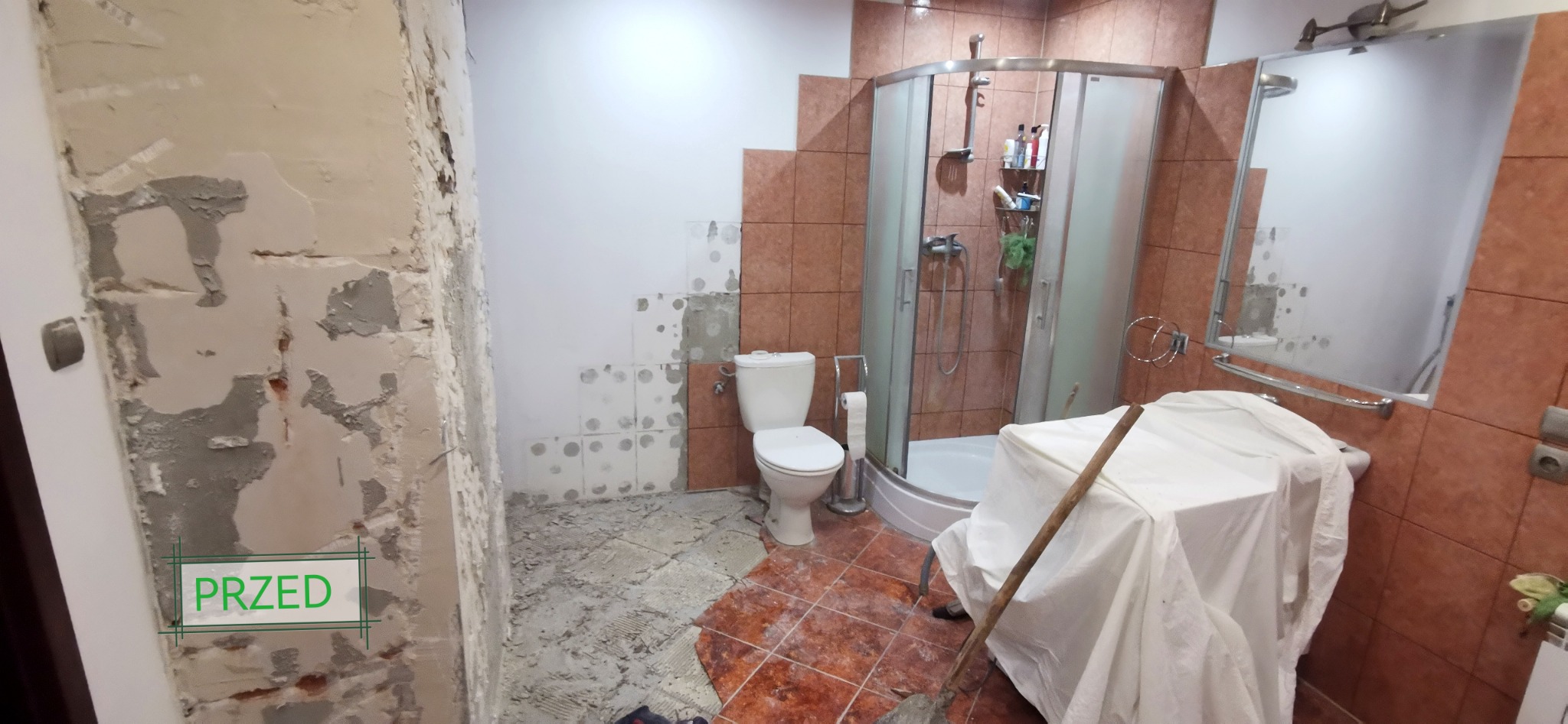 Łazienka w trakcie demontażu płytek i przygotowania ścian do dalszych prac, widoczny fragment ściany z napisem 'PRZED', częściowo usunięte płytki z podłogi i ścian, zakryta umywalka, kabina...