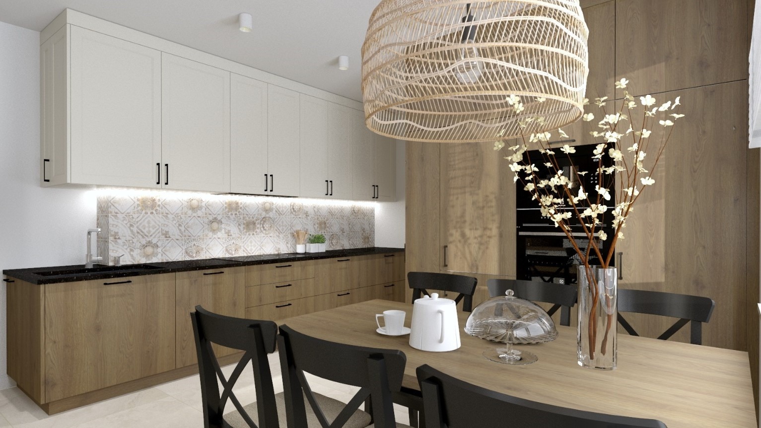 Jasna kuchnia z drewnianymi i białymi szafkami, czarnym blatem i wzorzystą ścianą nad nim. Nad stołem wisi pleciona lampa, a na stole stoi wazon z gałązkami, porcelana i szklany klosz.