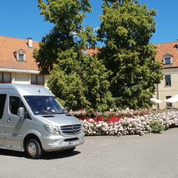Barbara Ślesicka - Srebrny bus Mercedes-Benz z przyciemnianymi szybami na tle dziedzińca z budynkami z czerwonymi dachami i kwitnącymi rabatami kwiatowymi.