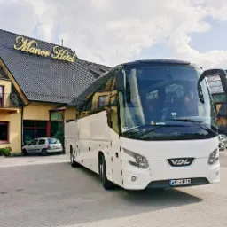Biały autokar VDL zaparkowany przed hotelem Manor, widoczne otwarte drzwi. Na pierwszym planie kostka brukowa.