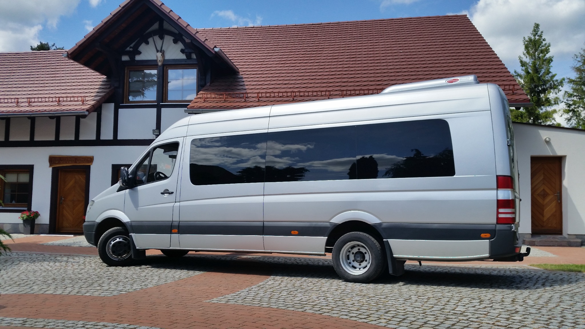 Srebrny bus z przyciemnianymi szybami zaparkowany przed budynkiem z czerwoną dachówką i białymi ścianami, odbicie chmur w oknach.