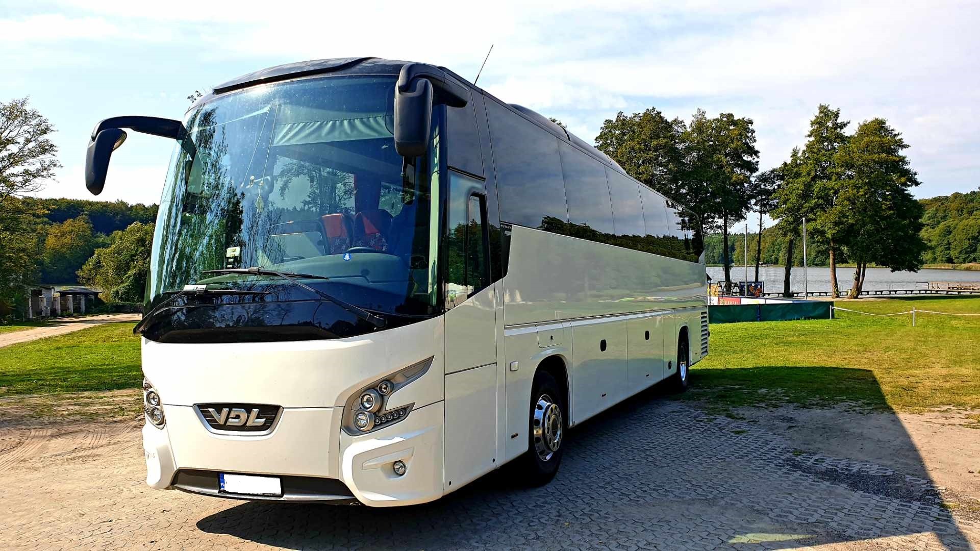 Biały autokar VDL zaparkowany na brukowanej powierzchni z widokiem na jezioro i las w tle, oświetlony słonecznym światłem.