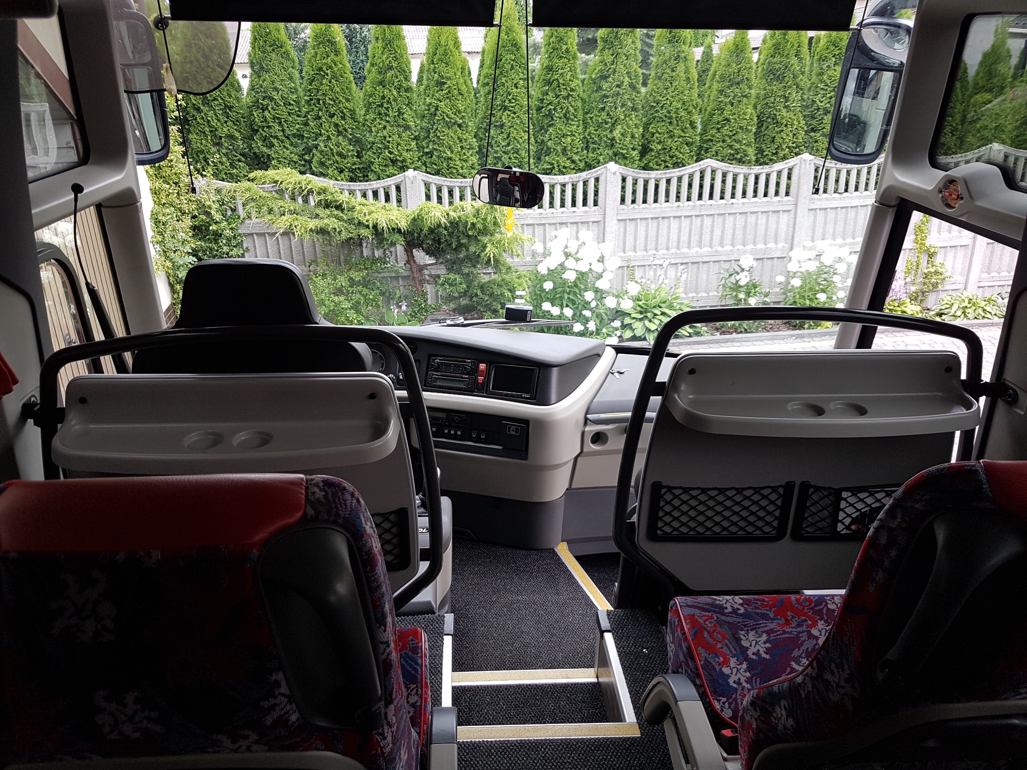 Wnętrze autobusu z widokiem na zielone tuje i białe kwiaty za oknem, fotele z wzorzystą tapicerką i stolikami.