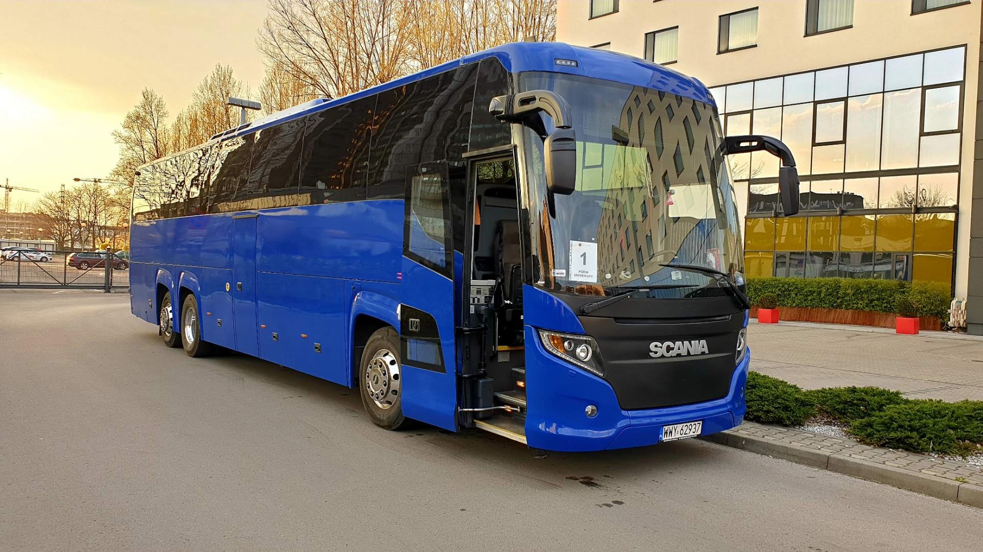 Niebieski autokar Scania z otwartymi drzwiami stoi przed budynkiem z refleksami zachodzącego słońca w oknach.