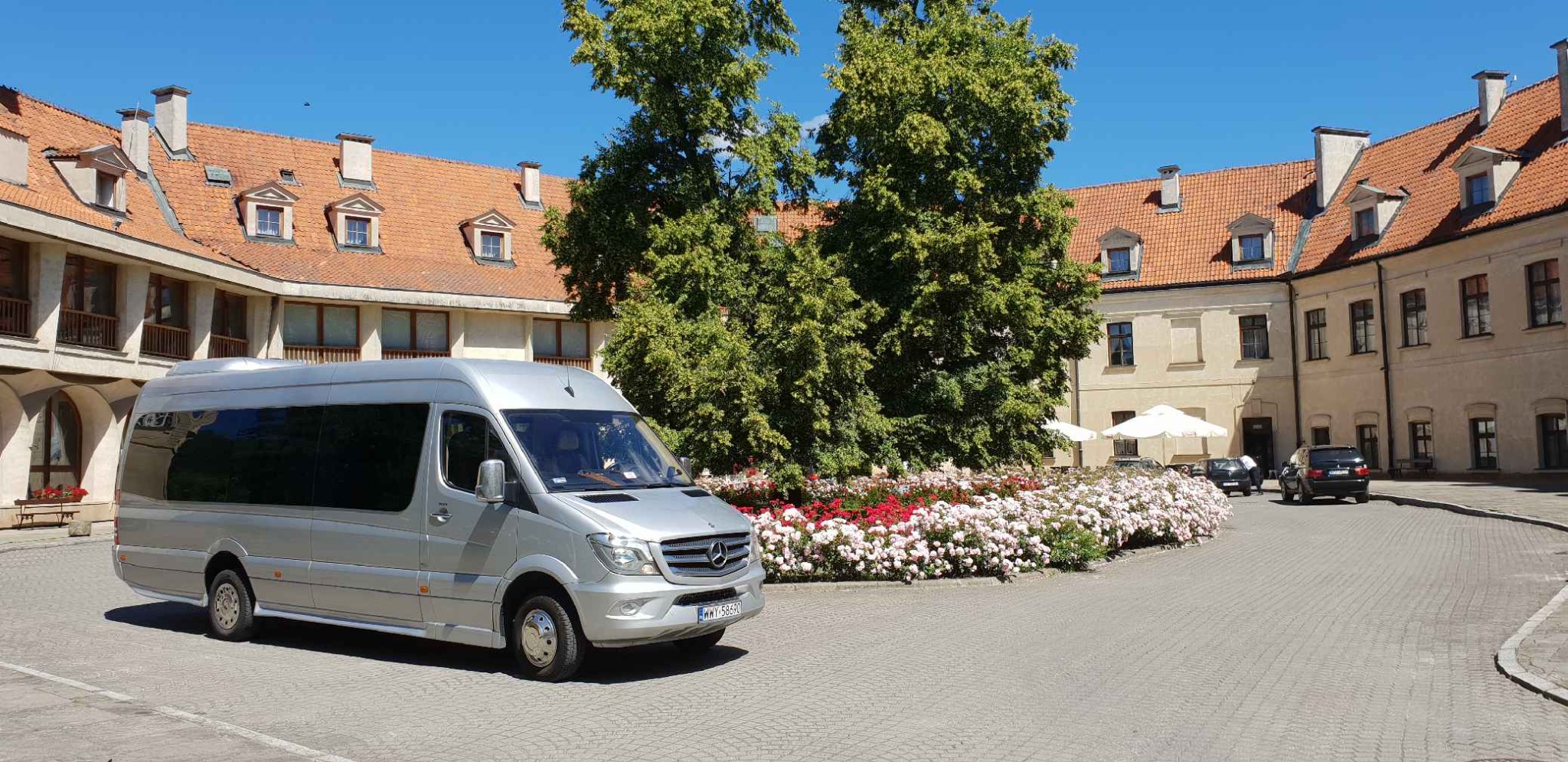 Srebrny bus Mercedes-Benz z przyciemnianymi szybami na tle dziedzińca z budynkami z czerwonymi dachami i kwitnącymi rabatami kwiatowymi.