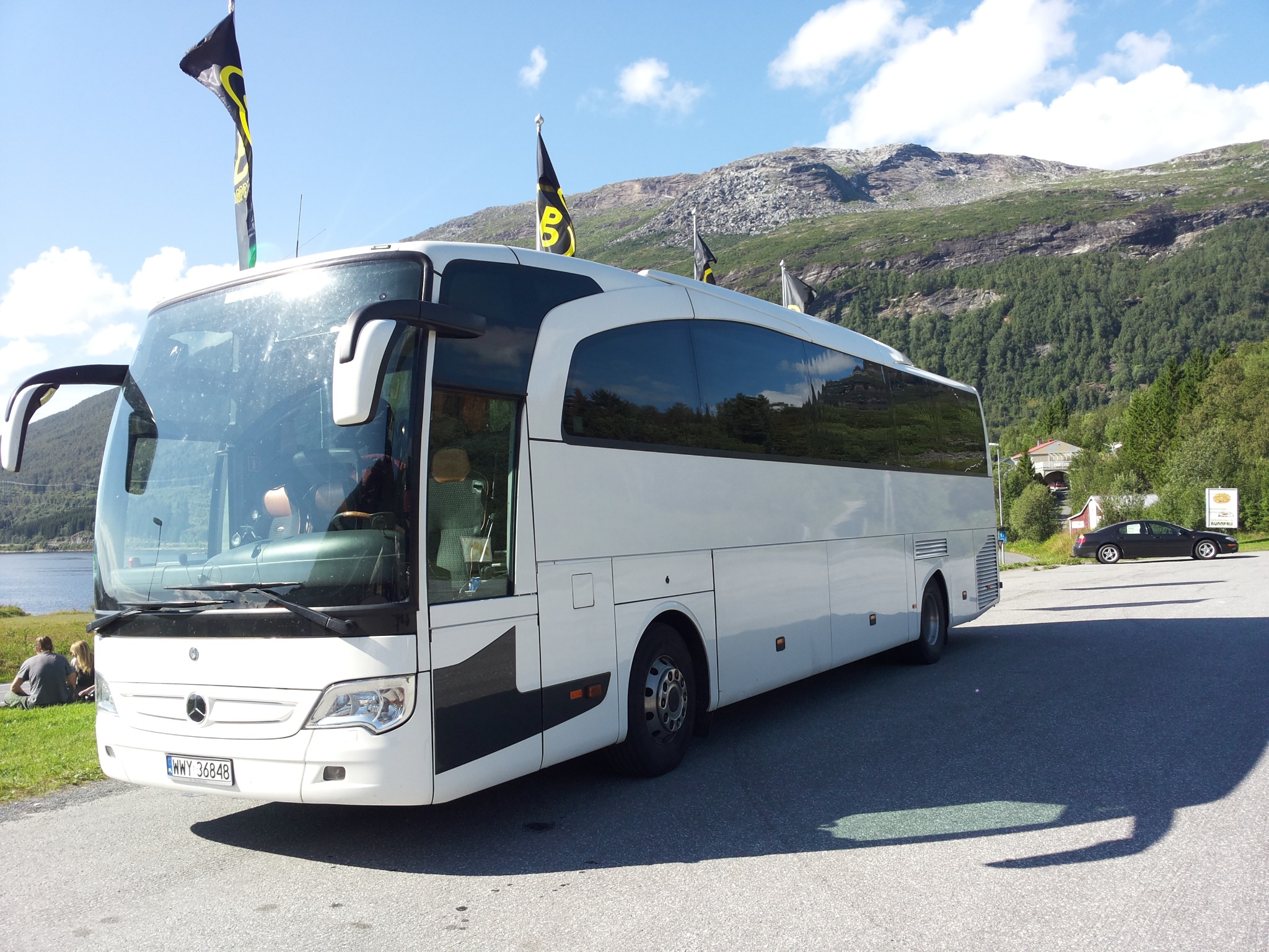 Biały autokar Mercedes z czarnymi flagami firmowymi na dachu, zaparkowany na tle górskiego krajobrazu i jeziora.