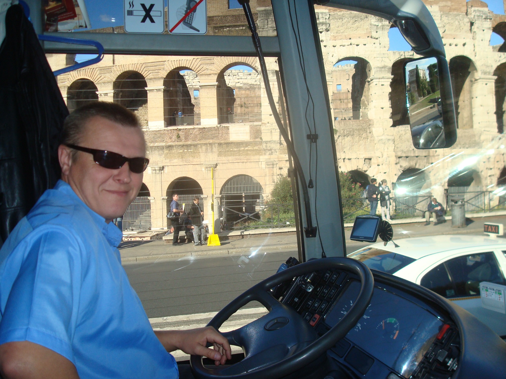 Kierowca autobusu w okularach przeciwsłonecznych uśmiecha się, na tle Koloseum w Rzymie, widoczne przez szybę pojazdu.