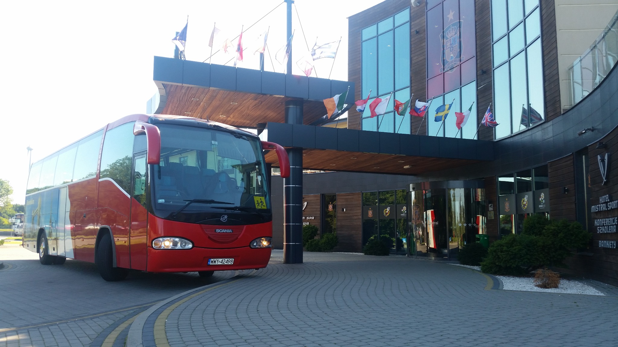 Czerwony autokar Scania z tablicą rejestracyjną WWY 42498 przed hotelem Mistral Sport z flagami różnych państw.