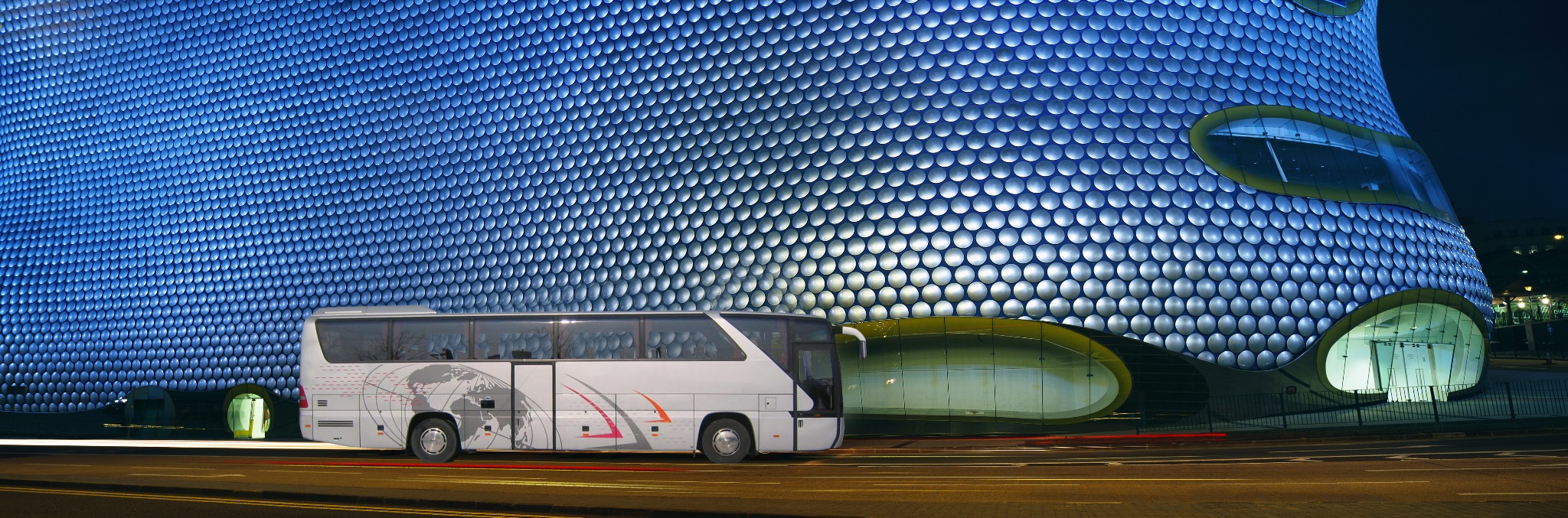 Autokar turystyczny z grafiką mapy świata zaparkowany przed nowoczesnym budynkiem o futurystycznej architekturze.
