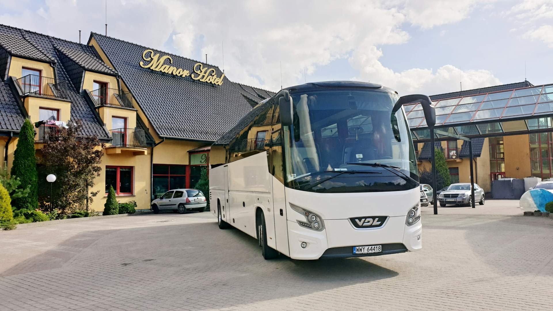 Biały autokar VDL zaparkowany przed hotelem Manor, widoczne otwarte drzwi. Na pierwszym planie kostka brukowa.