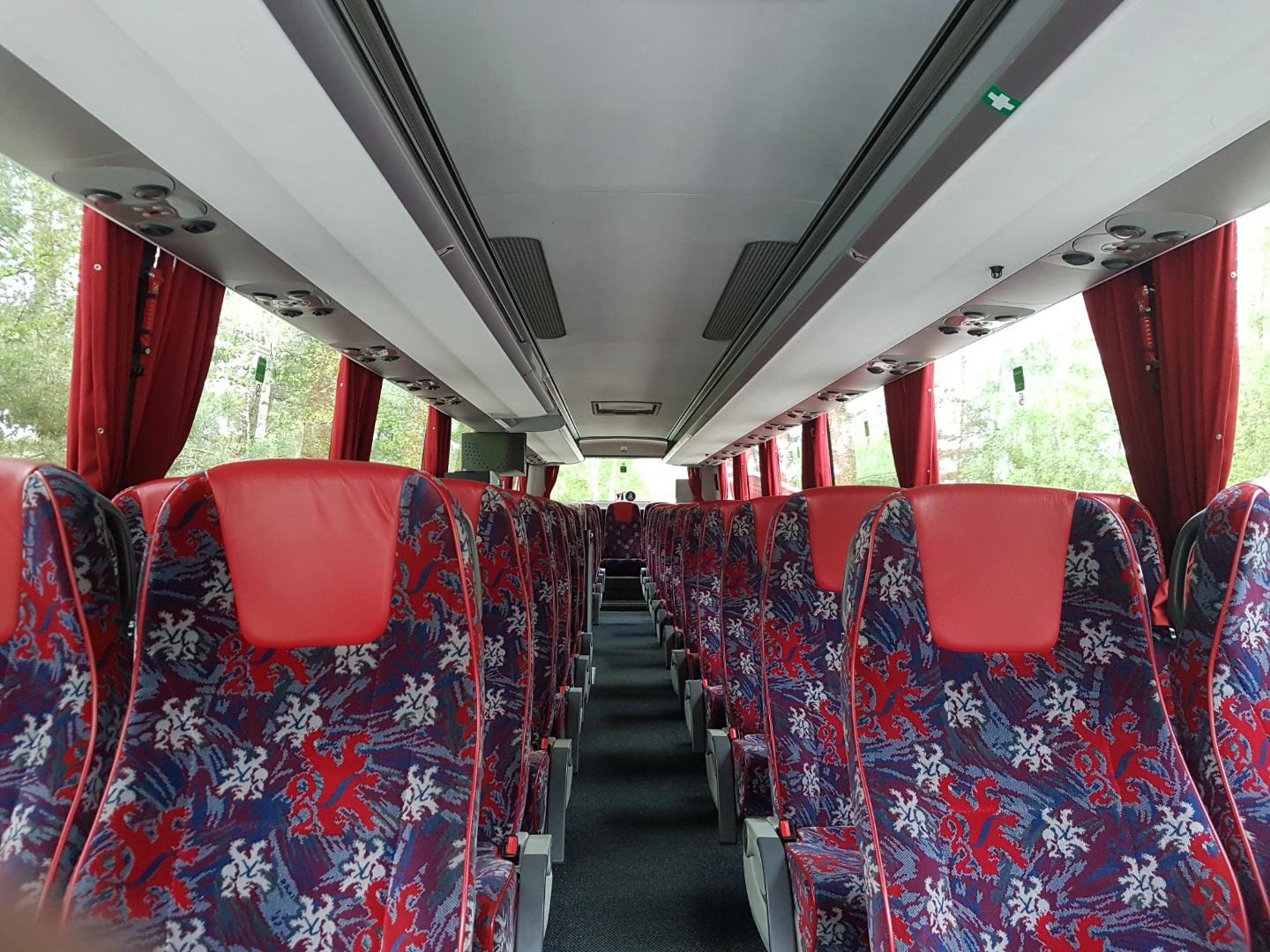 Wnętrze autobusu turystycznego z czerwonymi zasłonami i fotelami w kolorze czerwono-niebieskim z białym wzorem, widok z przodu kabiny pasażerskiej.