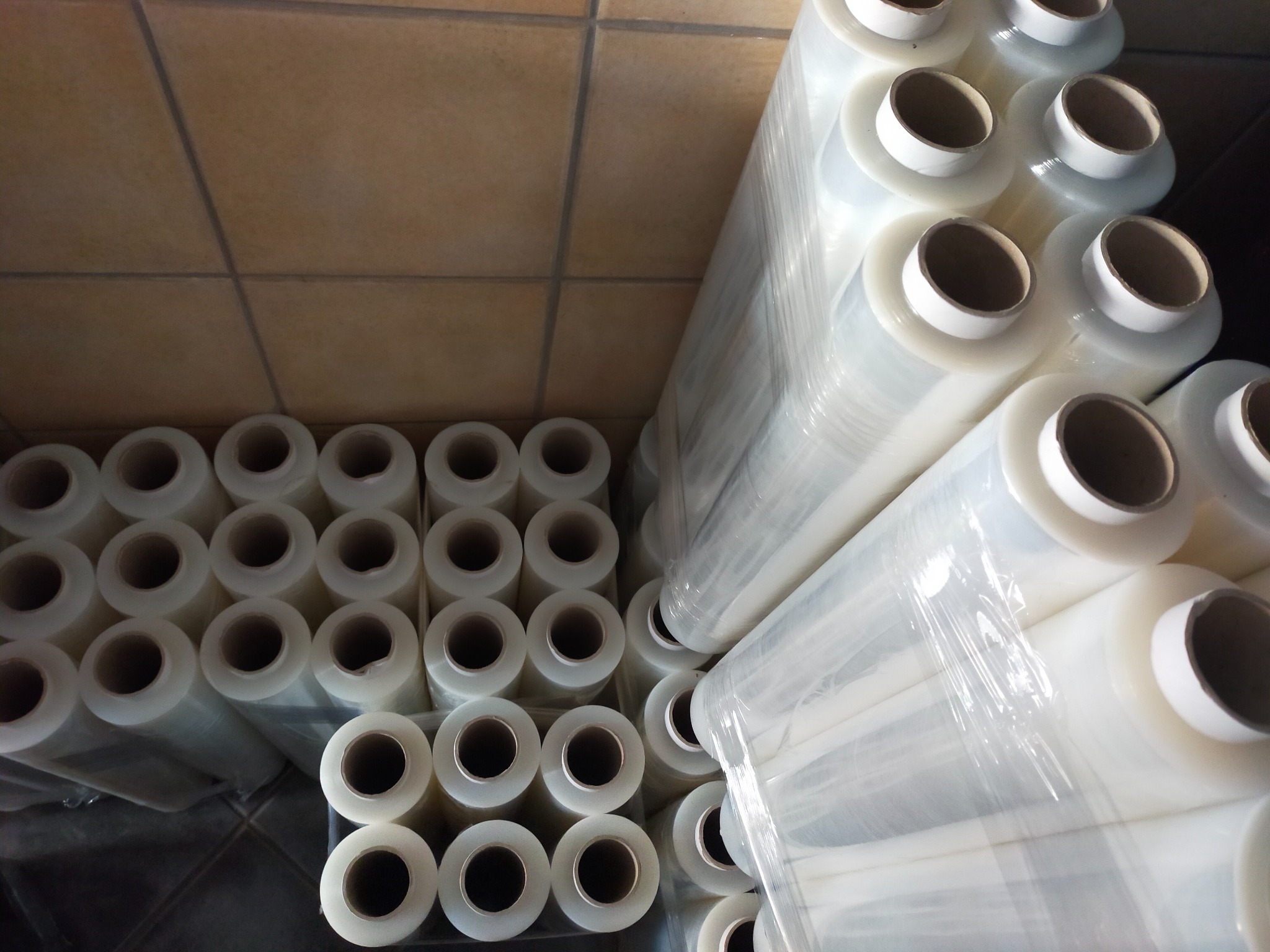 Stosy rolek bezbarwnej folii stretch 23 mikrony, ułożone na podłodze z płytek, widoczne z góry.