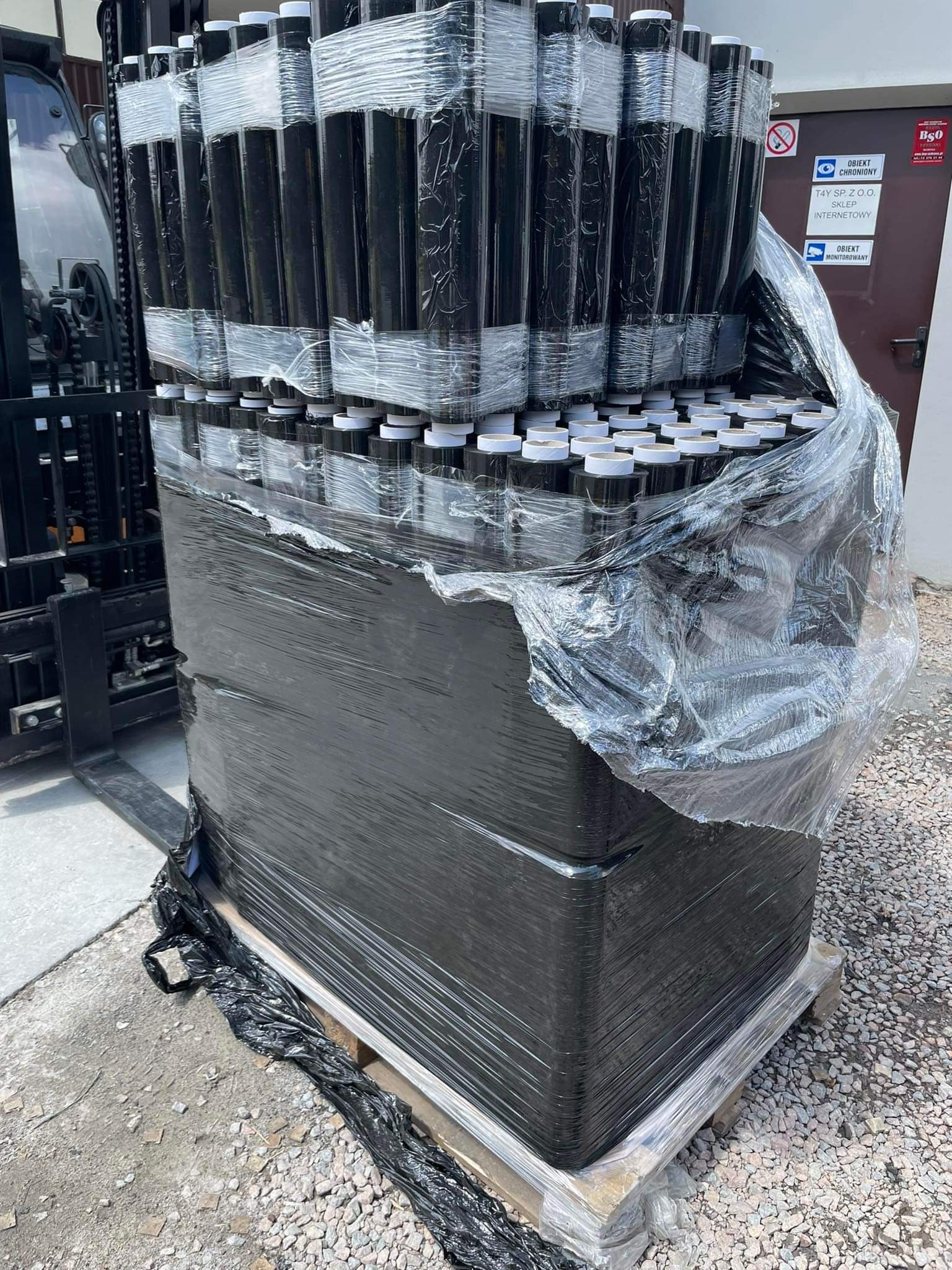 Paleta z czarnymi, cylindrycznymi opakowaniami z białymi wieczkami, owinięta folią stretch, transportowana wózkiem widłowym na zewnątrz budynku.