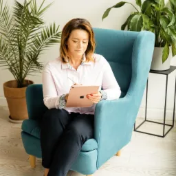Kobieta w jasnej koszuli i ciemnych spodniach siedzi w turkusowym fotelu z wysokim oparciem, trzymając w rękach tablet i rysik, obok doniczkowe rośliny i minimalistyczny stolik.