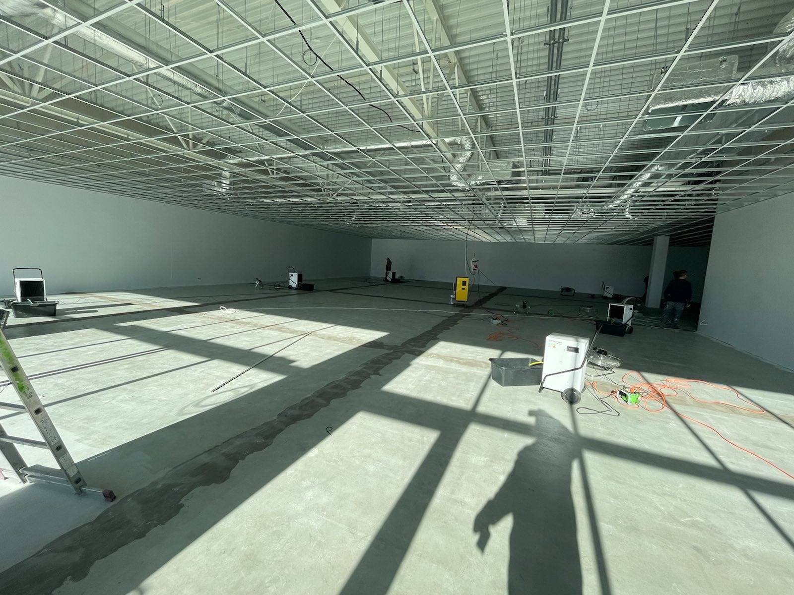 Wnętrze hali w trakcie osuszania: widoczne urządzenia osuszające, metalowa konstrukcja sufitu, jasne ściany, długie cienie padające na podłogę. Na podłodze widoczne ślady po zalaniu.