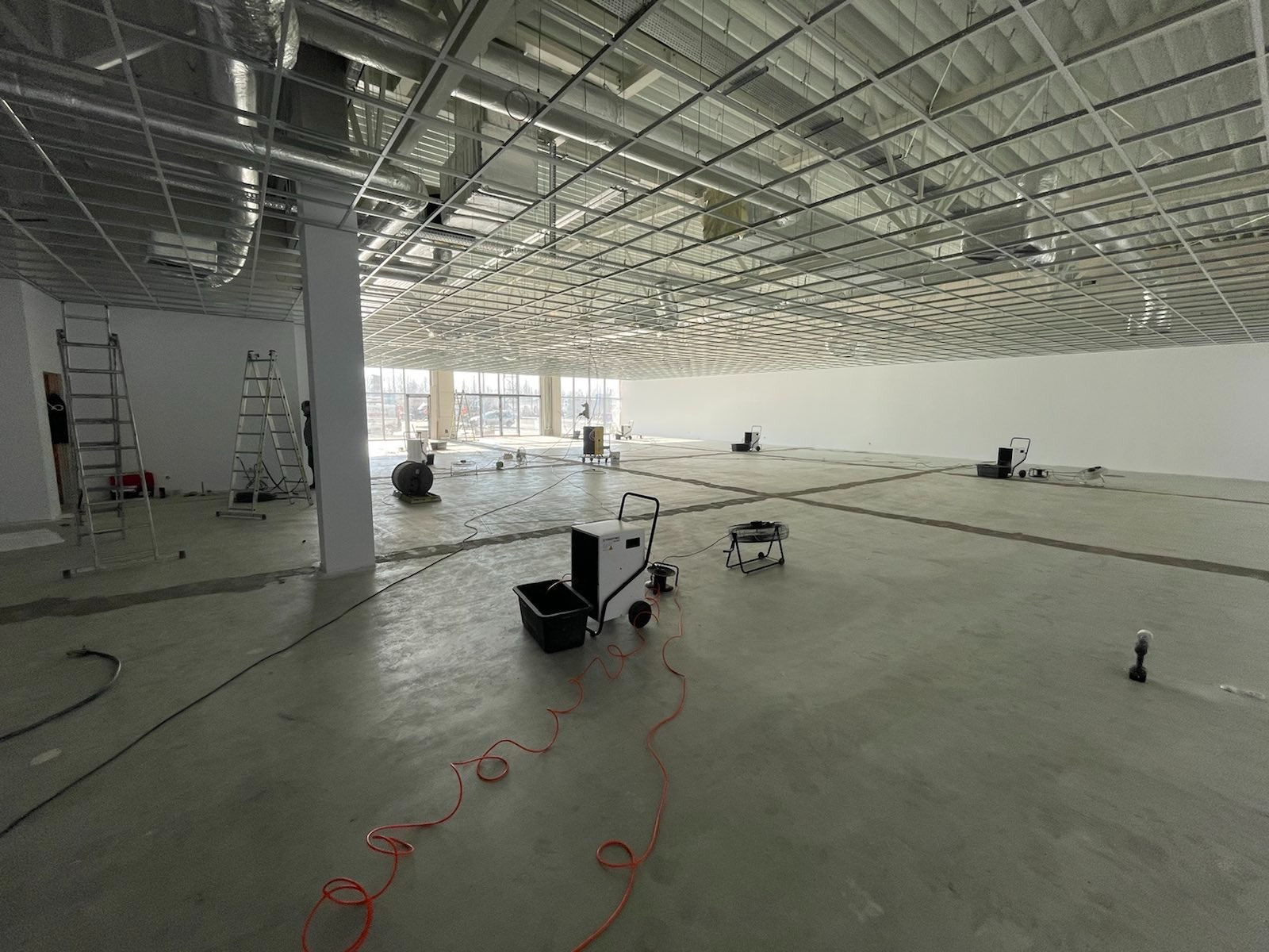 Rozległe wnętrze w trakcie osuszania posadzki, widoczne osuszacze kondensacyjne, wentylatory i drabiny, surowy beton i metalowa konstrukcja sufitu podwieszanego, naturalne światło wpadające przez...