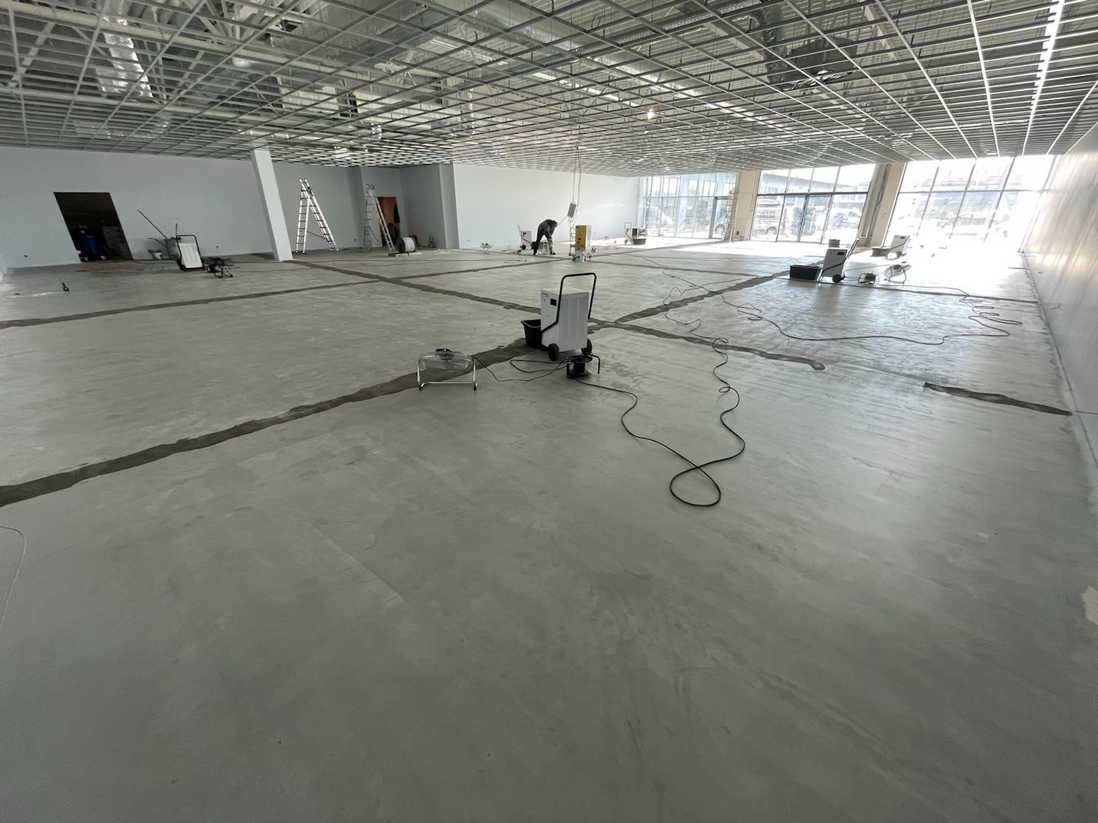 Rozległa, szara betonowa posadzka w trakcie schnięcia w nowym budynku, z widocznymi czarnymi liniami dylatacyjnymi, osuszaczem kondensacyjnym, wentylatorem podłogowym i pracownikiem w oddali.
