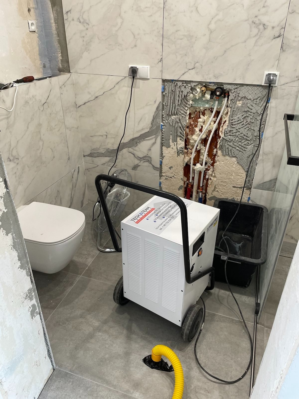 Wnętrze łazienki w trakcie remontu, widoczny osuszacz powietrza firmy Tech Team, podłączony do żółtego węża, obok toaleta i ściana z odsłoniętymi rurami i instalacjami.
