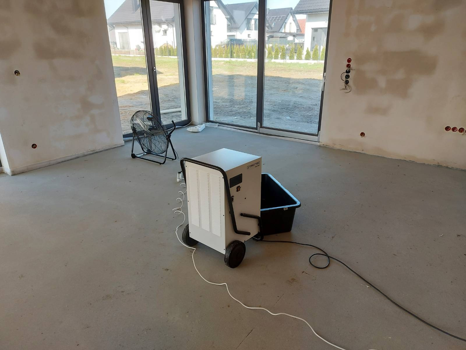 Przenośny osuszacz powietrza Trotec z czarnym zbiornikiem na wodę w surowym wnętrzu z oknami i wentylatorem, widoczne ślady wilgoci na ścianach.