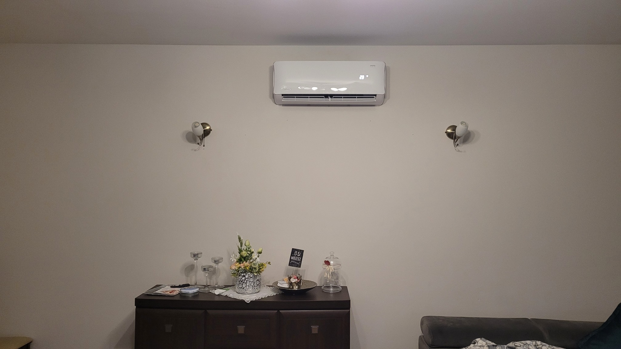 Biały klimatyzator ścienny zamontowany w pokoju nad ciemną komodą z dekoracjami, oświetlony dwoma kinkietami