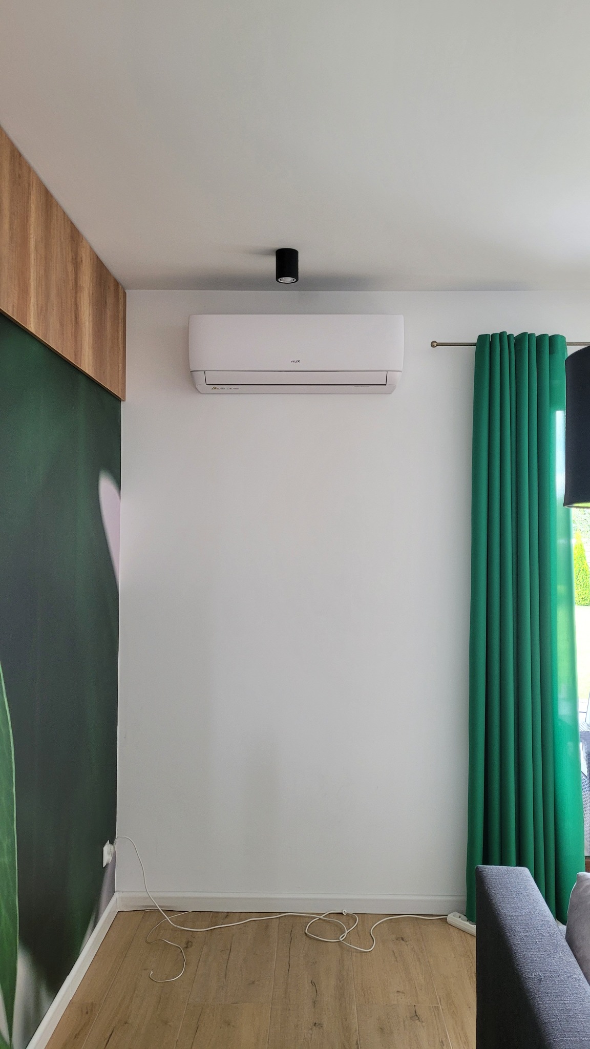 Biały klimatyzator marki AUX zamontowany na ścianie w minimalistycznym salonie z zielonymi zasłonami i drewnianą podłogą, z widocznym przewodem zasilającym przy podłodze.