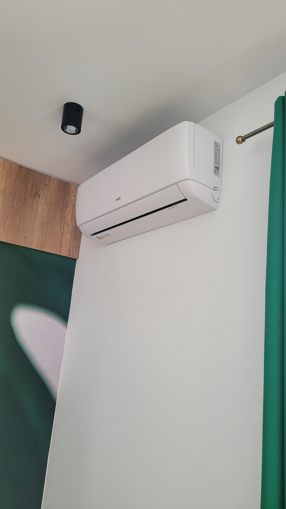 Biała jednostka klimatyzacyjna marki AUX zamontowana na białej ścianie w pomieszczeniu z fragmentem drewnianej boazerii i zielonej zasłony.