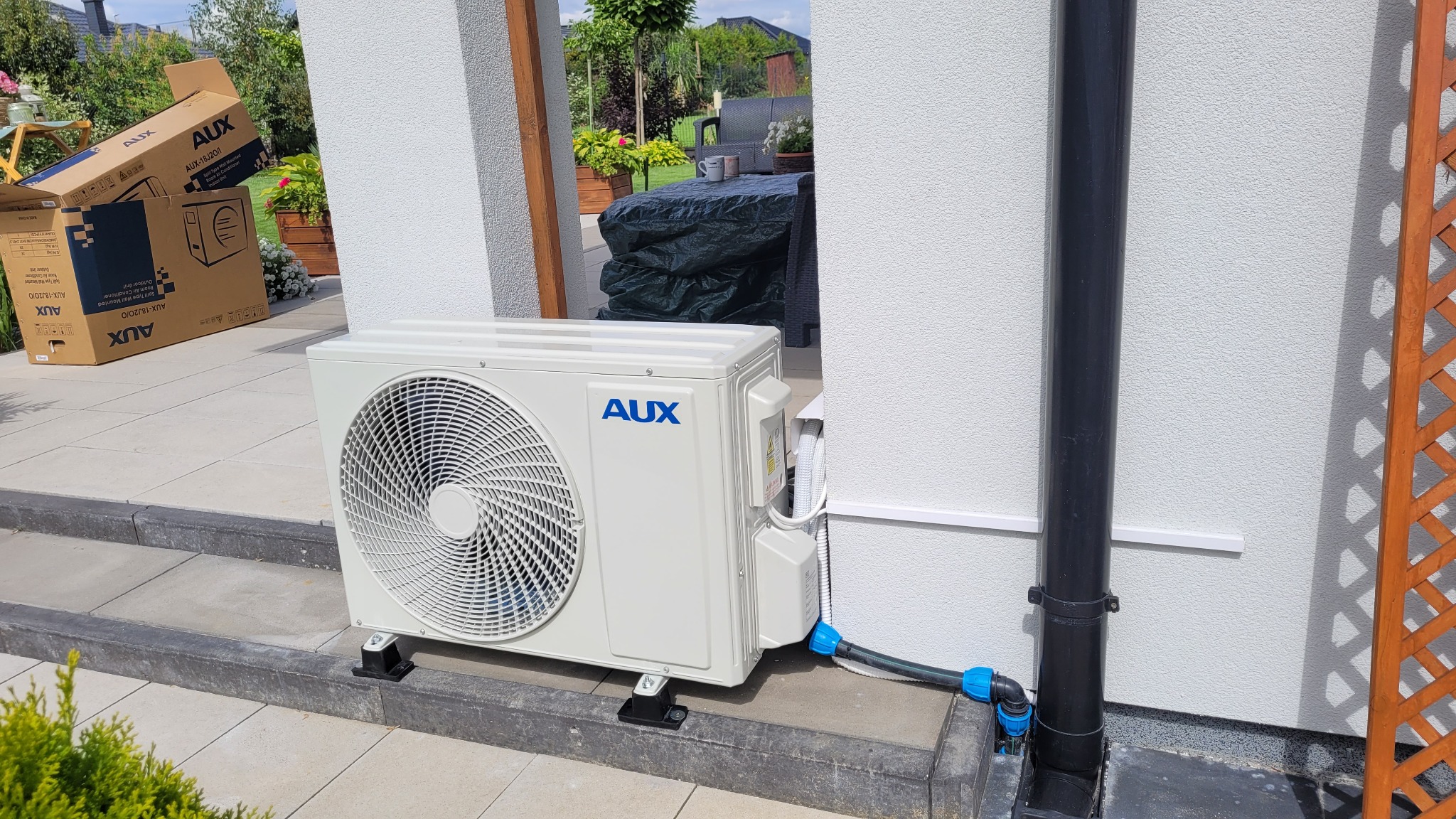 Zewnętrzna jednostka klimatyzacji AUX zamontowana na betonowym podeście przy ścianie domu, z widoczną instalacją odprowadzającą skropliny i kartonem po urządzeniu w tle.