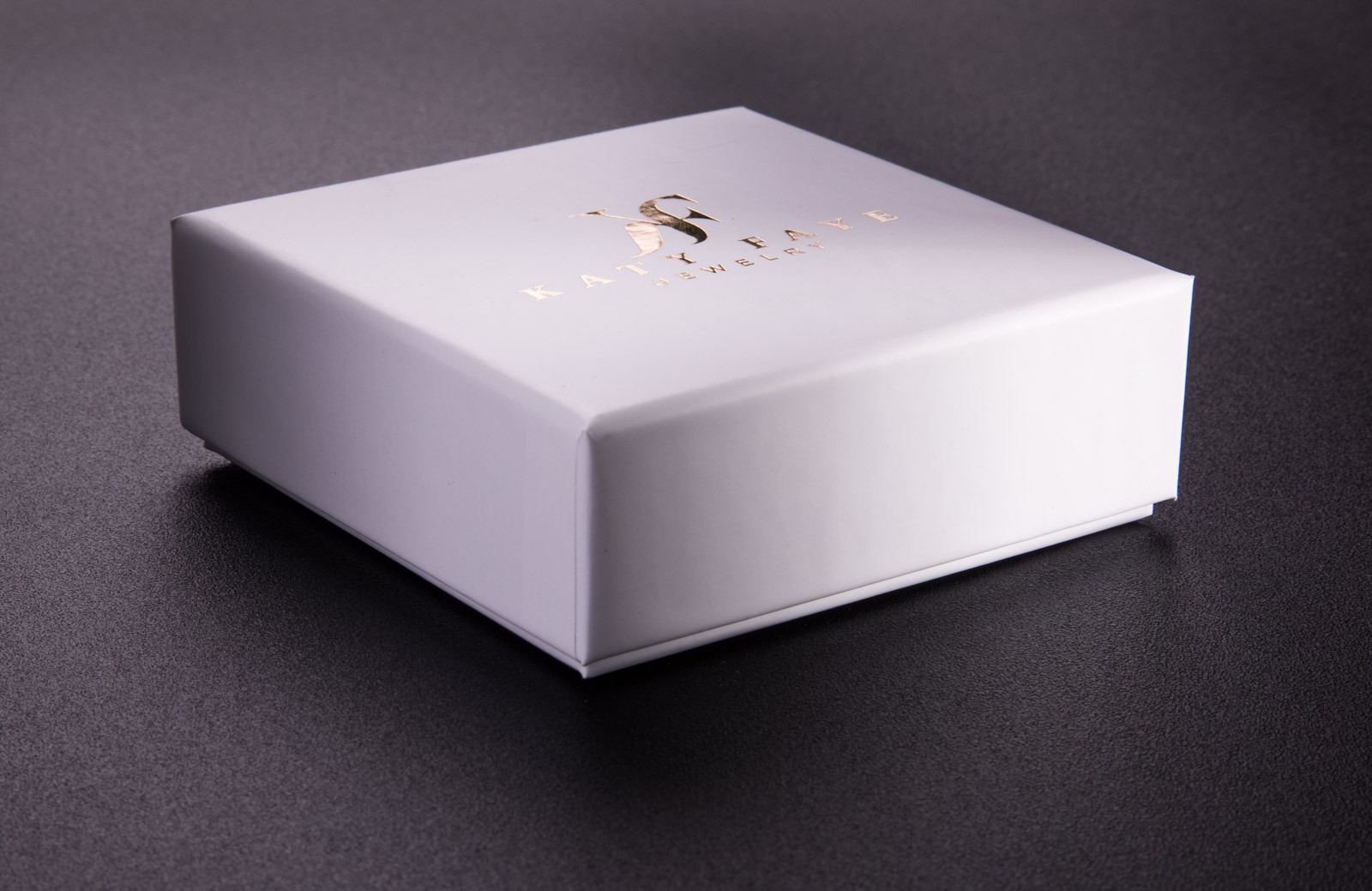 Białe, kwadratowe pudełko z logo w kolorze złotym, umieszczone na ciemnoszarym tle. Widoczny subtelny cień.