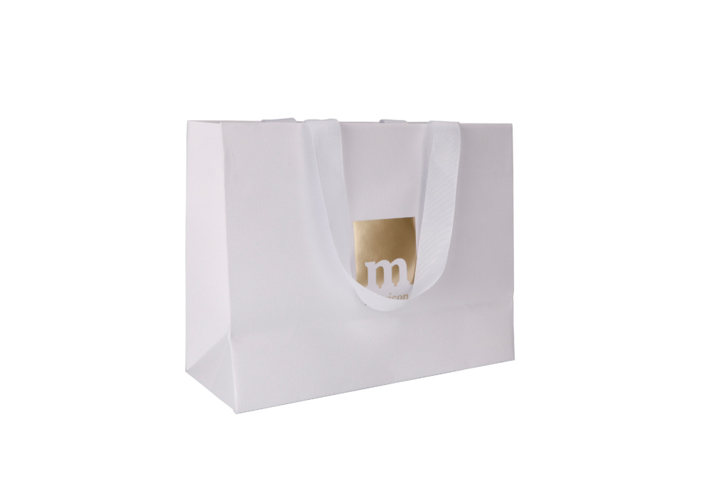 Biała torba papierowa z satynowymi uchwytami i złotym logo firmy, ustawiona na białym tle.