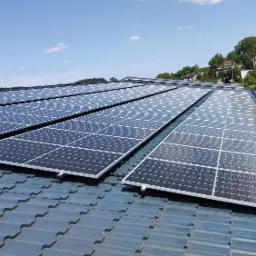 Instalacja fotowoltaiczna na dachu krytym szarą dachówką, widok z dołu pod kątem, w tle zabudowania i zieleń.