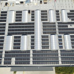 PROEKO OZE SPÓŁKA Z OGRANICZONĄ ODPOWIEDZIALNOŚCIĄ - Widok z góry na rozległą instalację paneli słonecznych na dachu budynku przemysłowego, z elementami wentylacji i zieleni w tle. Układ paneli w regularnych rzędach.