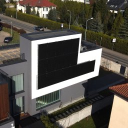 PROEKO OZE SPÓŁKA Z OGRANICZONĄ ODPOWIEDZIALNOŚCIĄ - Nowoczesny dom z panelami słonecznymi na elewacji, widok z góry. Minimalistyczna architektura, szaro-biała kolorystyka, otoczenie zieleni i zabudowa miejska w tle.