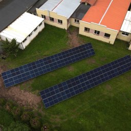 PROEKO OZE SPÓŁKA Z OGRANICZONĄ ODPOWIEDZIALNOŚCIĄ - Widok z drona na dwa rzędy paneli fotowoltaicznych na trawiastym terenie, obok budynków z różnymi pokryciami dachowymi. Ujęcie z góry, równomierne oświetlenie.