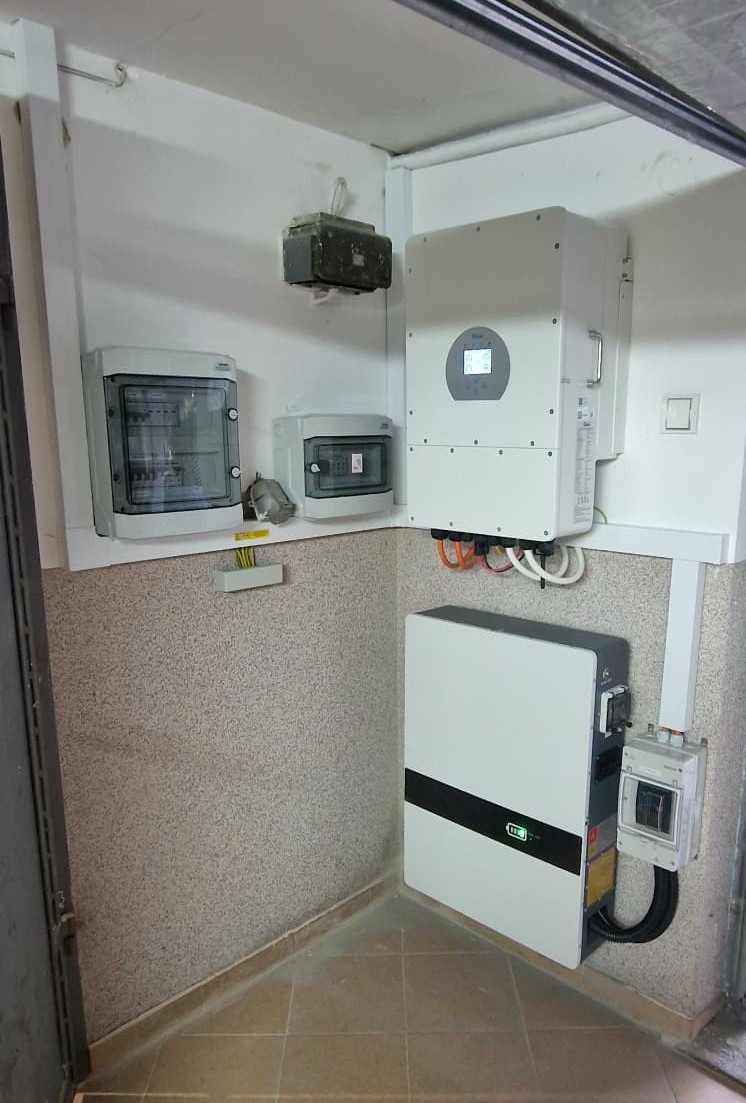 Wewnętrzna instalacja fotowoltaiczna: biały inwerter z wyświetlaczem i akumulator energii na ścianie w rogu pomieszczenia, widoczne rozdzielnice elektryczne i okablowanie.