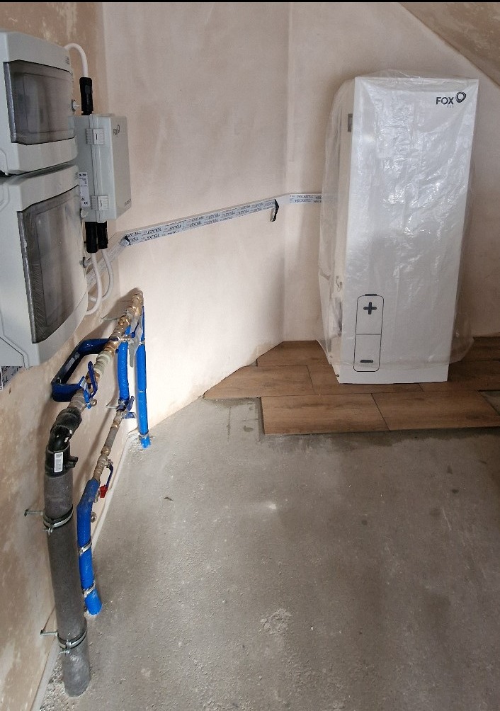 Wnętrze pomieszczenia w trakcie montażu instalacji: widoczne rozdzielnice elektryczne, rury instalacyjne w niebieskiej izolacji oraz biały akumulator energii w folii ochronnej, częściowo ułożona...