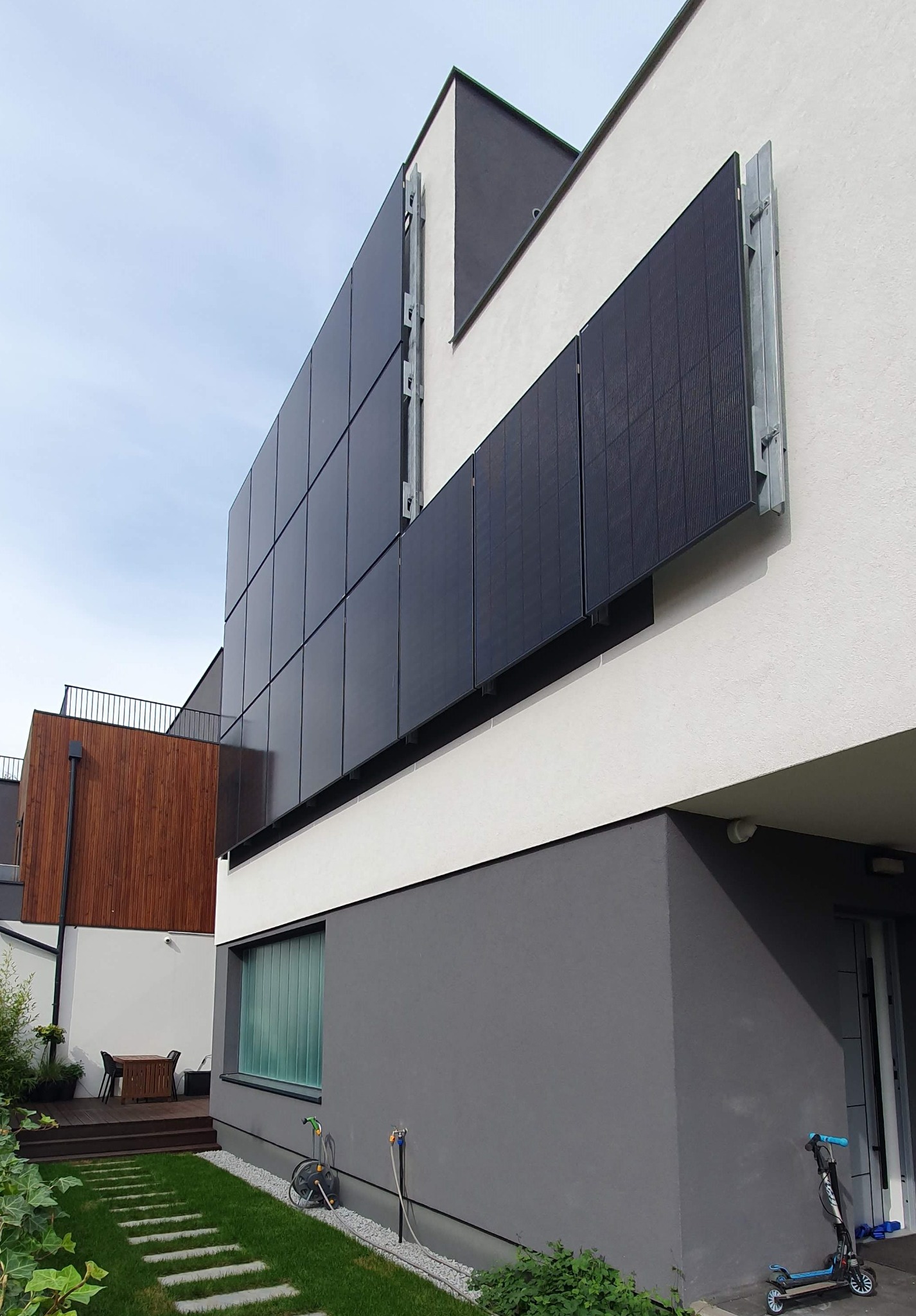 Nowoczesny dom z panelami fotowoltaicznymi zamontowanymi na fasadzie, widoczne metalowe uchwyty mocujące, trawnik z betonowymi płytami, hulajnoga oparta o ścianę.