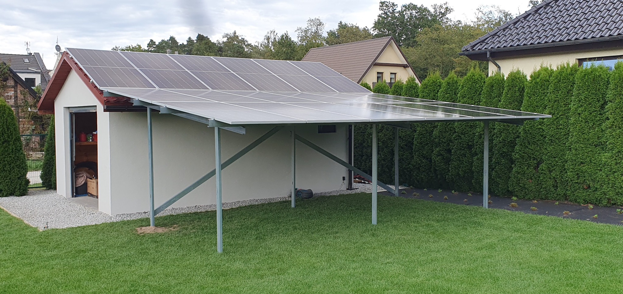 Instalacja fotowoltaiczna na dachu garażu z metalową konstrukcją wsporczą, widoczny robot koszący na trawniku i rząd tuj w tle.