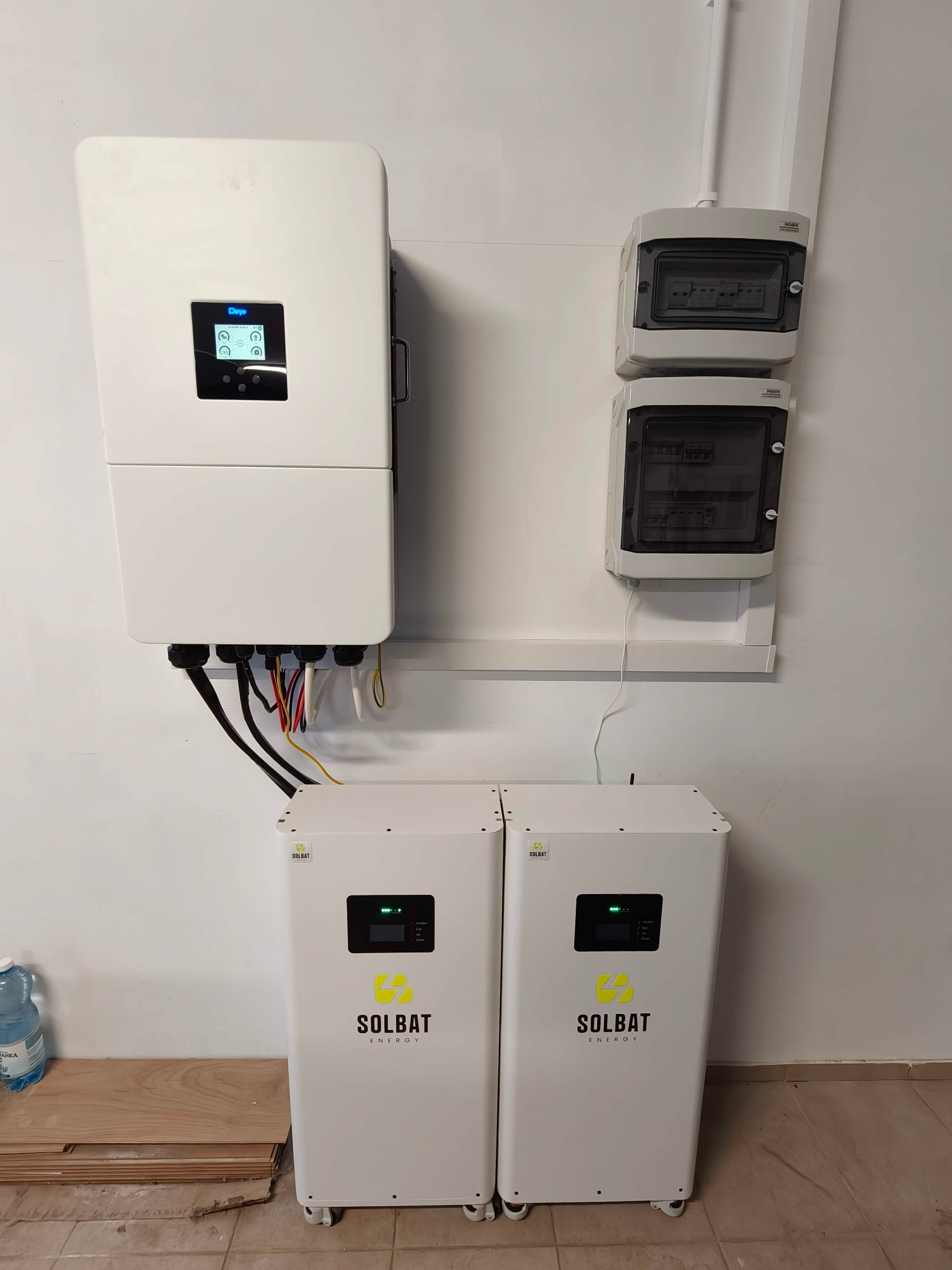 Biały inwerter i dwa akumulatory Solbat Energy zamontowane na ścianie, z widocznymi przewodami i skrzynkami elektrycznymi. U dołu leżą panele podłogowe i butelka wody.