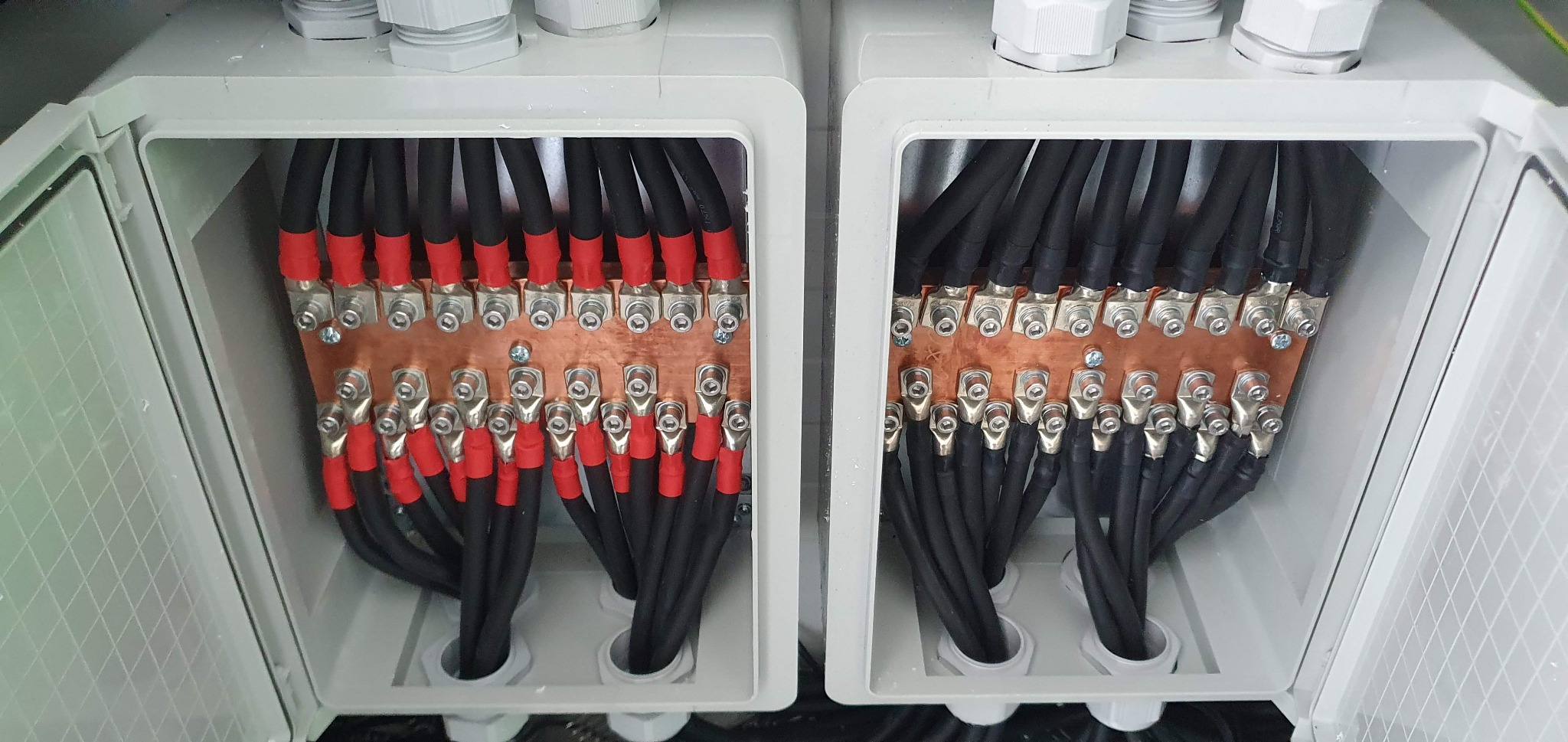 Dwa otwarte panele elektryczne z licznymi czarnymi kablami zakończonymi czerwonymi i srebrnymi końcówkami, podłączonymi do miedzianych szyn zbiorczych.