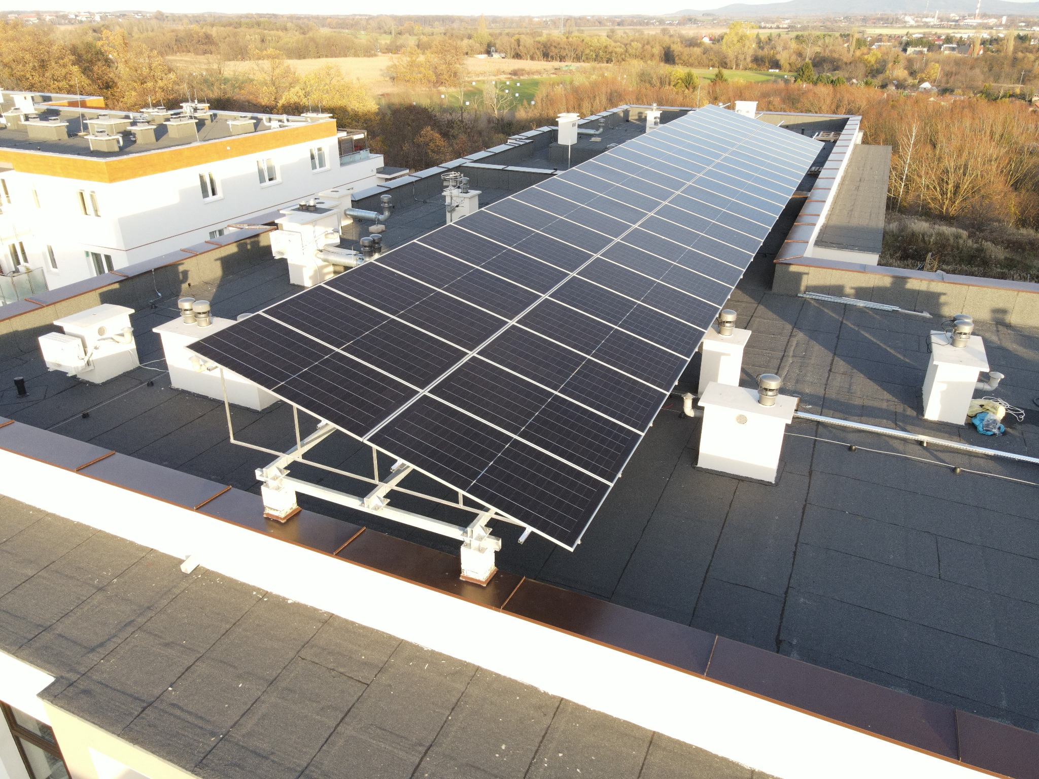 Panel słoneczny na dachu budynku z widokiem na drzewa w oddali. Instalacja fotowoltaiczna na płaskim dachu z elementami montażowymi. Widok z góry na system paneli.