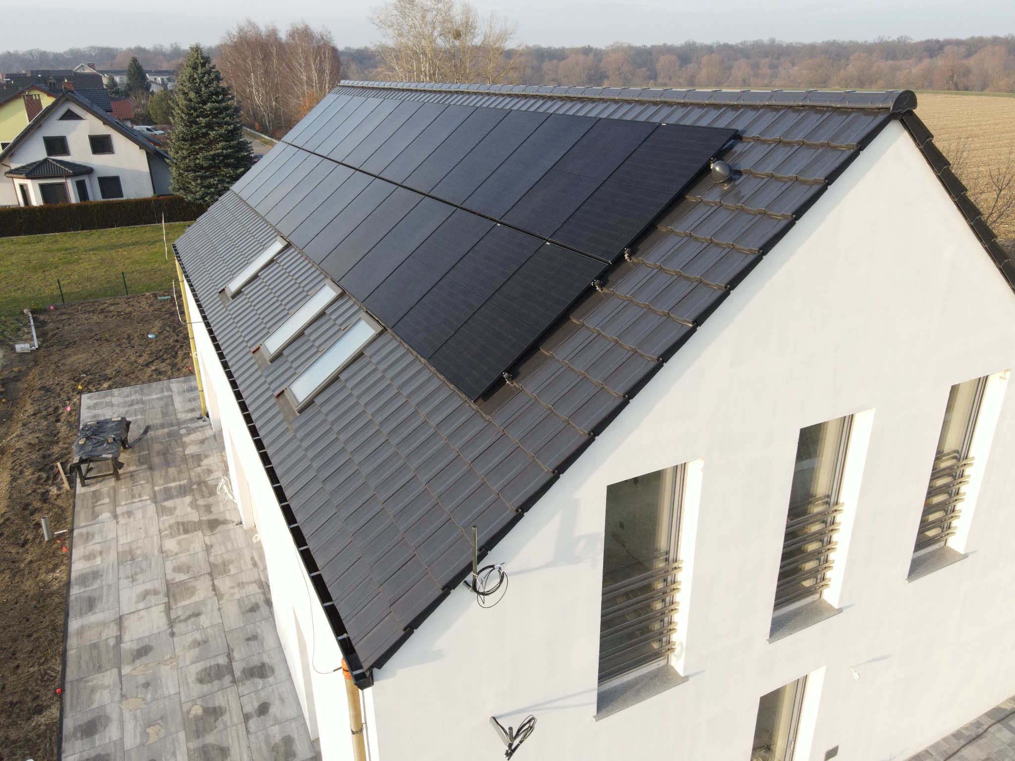 Czarny panel słoneczny na dachu domu z szarą dachówką i oknami dachowymi. Biała elewacja, kostka brukowa na podwórku. Widok z góry.