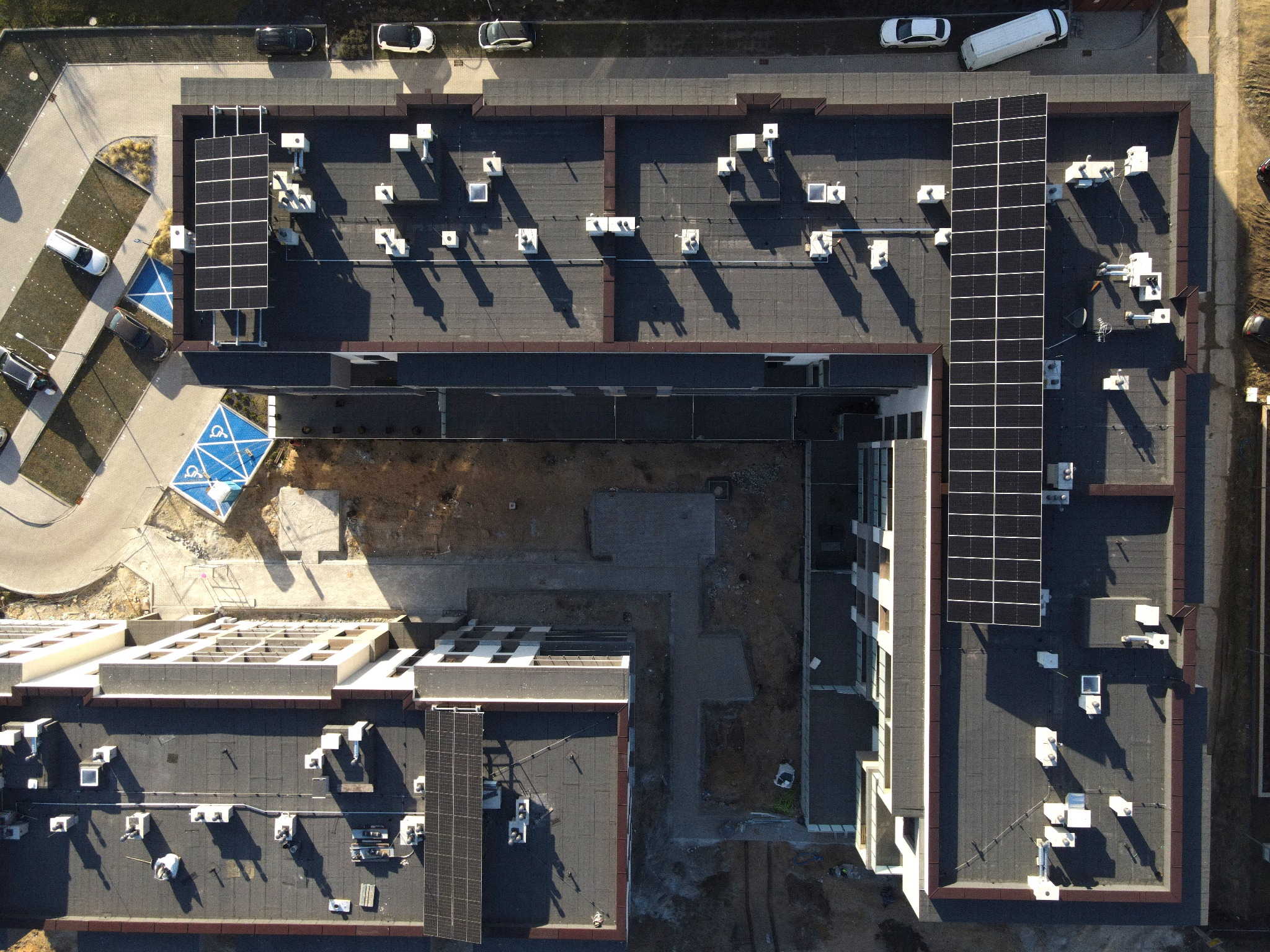 Widok z drona na dachy budynków z panelami słonecznymi i systemami wentylacji. Ciemna dachówka, jasne elementy wentylacyjne, panele w regularnych rzędach. Parking i zieleń w tle.