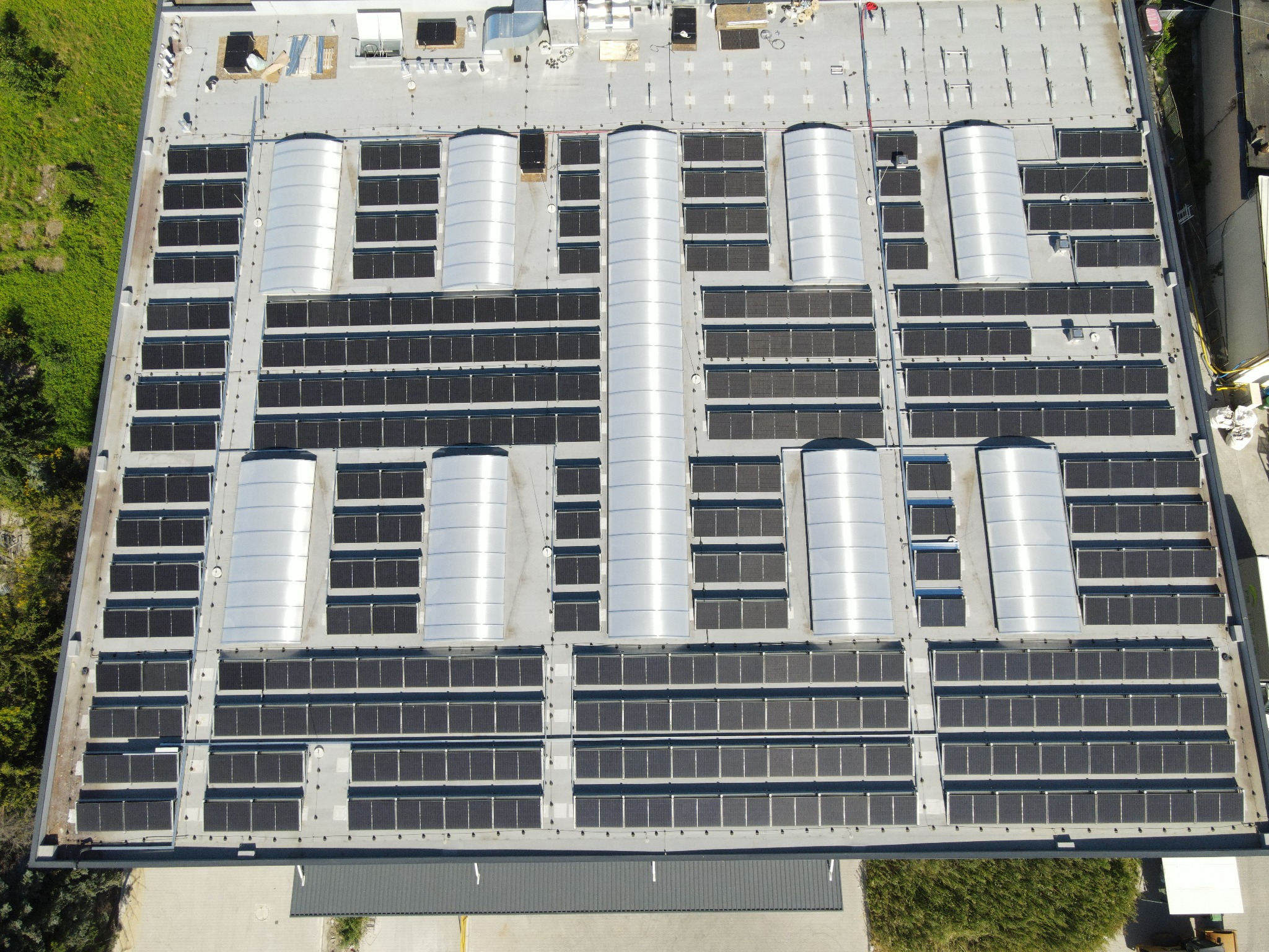 Widok z góry na rozległą instalację paneli słonecznych na dachu budynku przemysłowego, z elementami wentylacji i zieleni w tle. Układ paneli w regularnych rzędach.