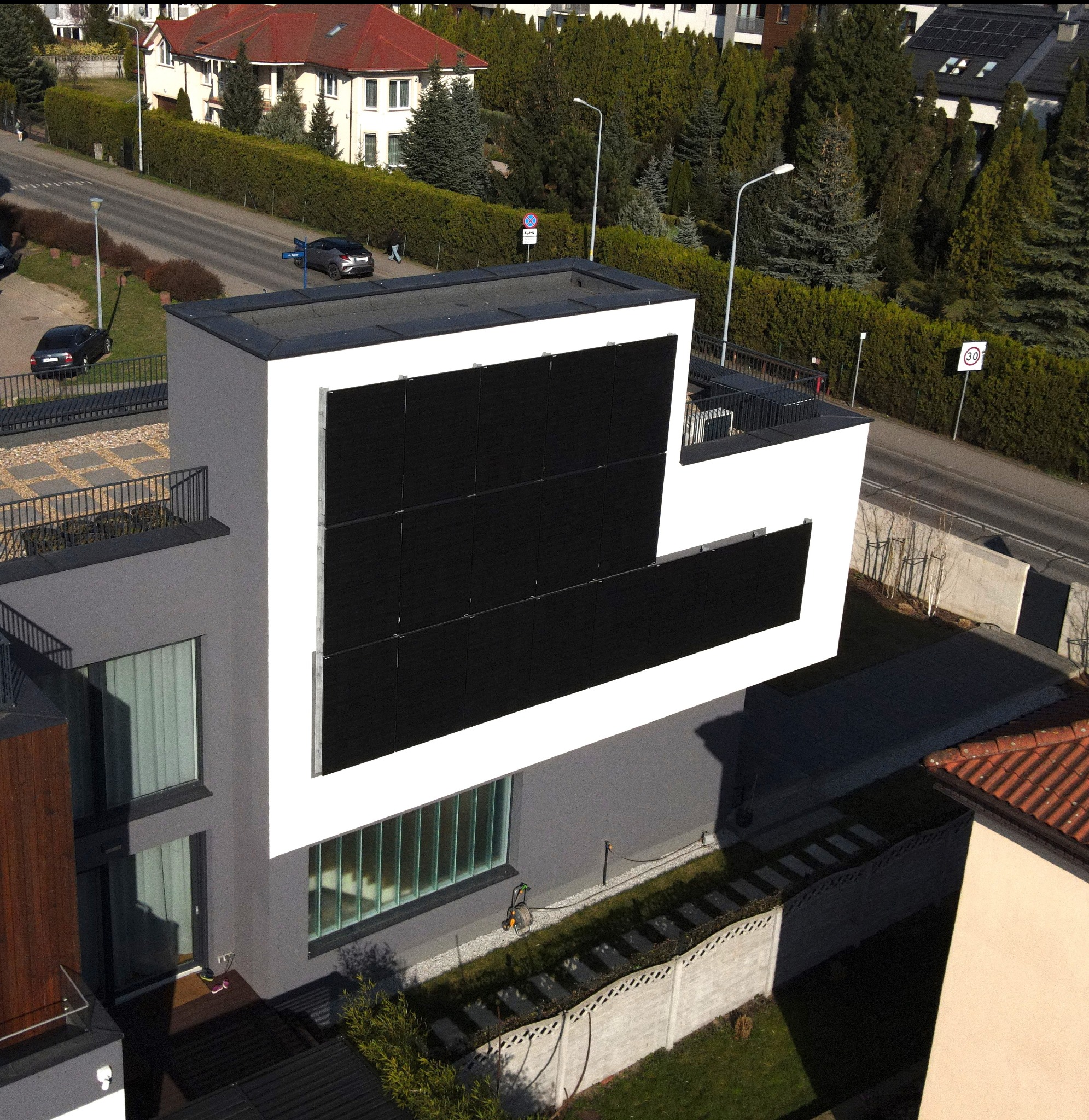 Nowoczesny dom z panelami słonecznymi na elewacji, widok z góry. Minimalistyczna architektura, szaro-biała kolorystyka, otoczenie zieleni i zabudowa miejska w tle.