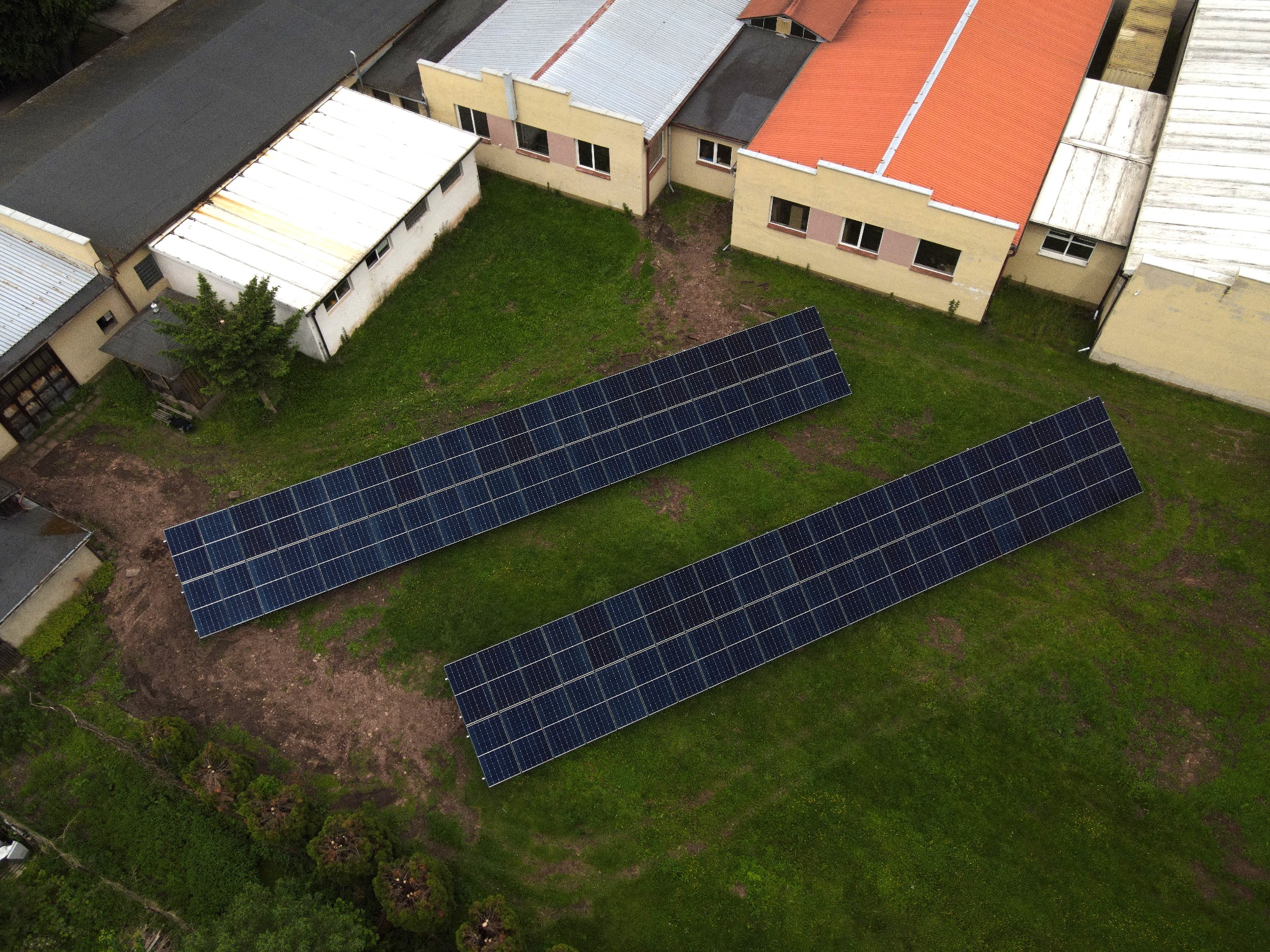 Widok z drona na dwa rzędy paneli fotowoltaicznych na trawiastym terenie, obok budynków z różnymi pokryciami dachowymi. Ujęcie z góry, równomierne oświetlenie.