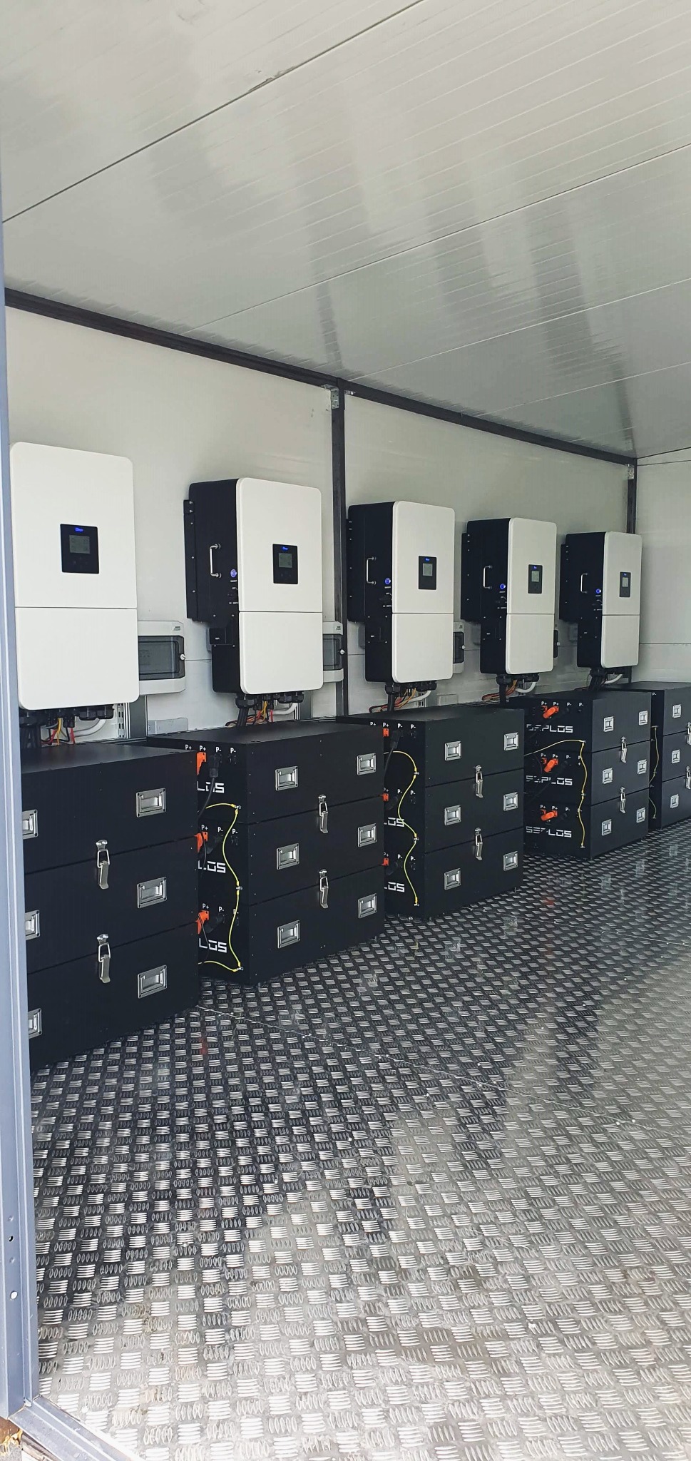 Rząd białych inwerterów i czarnych akumulatorów w metalowej obudowie z ryflowaną podłogą, tworzących system zasilania awaryjnego lub magazyn energii.