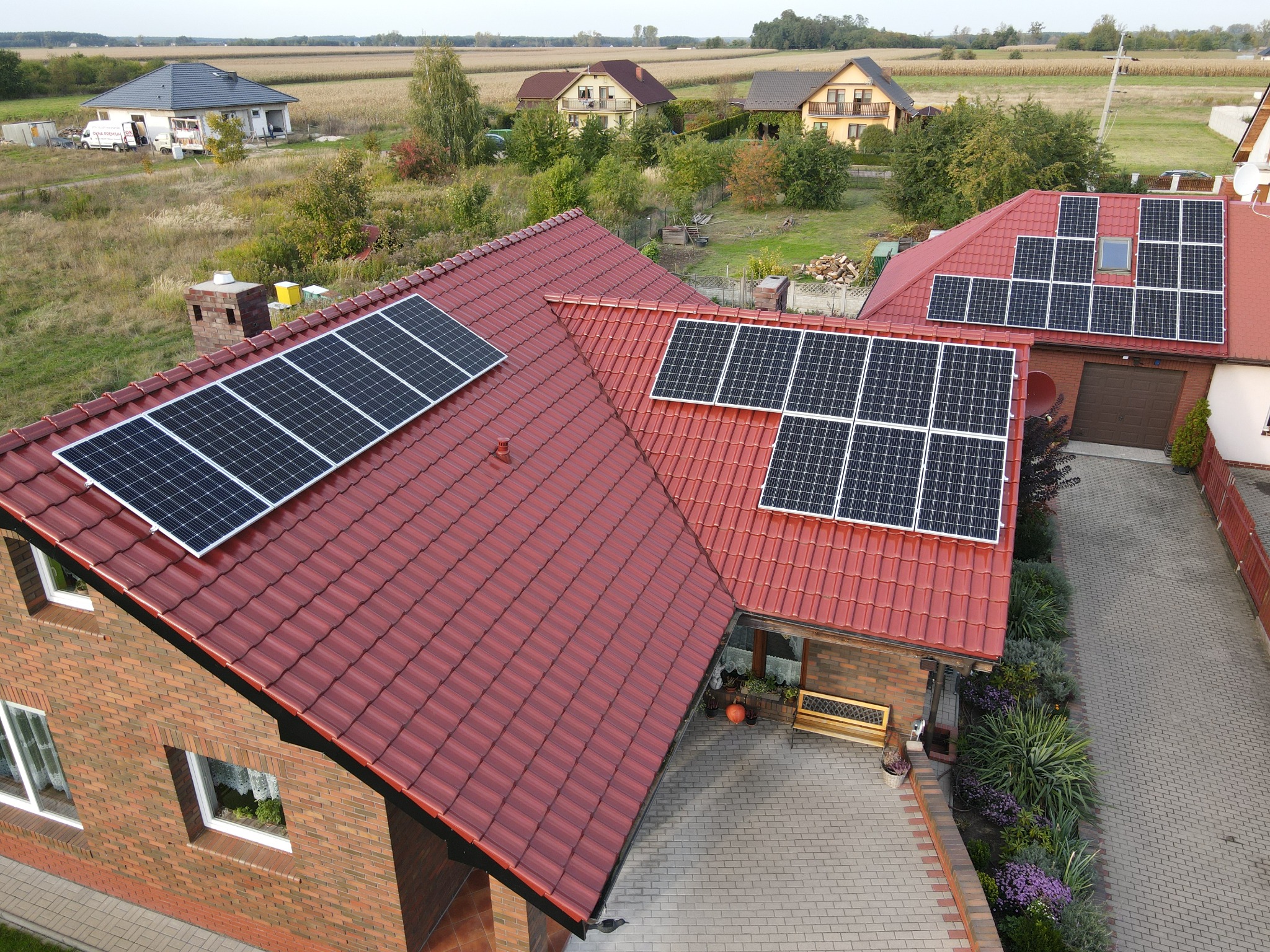 Czerwony dach z dachówki ceramicznej z panelami fotowoltaicznymi na dwóch budynkach mieszkalnych, widok z góry na podjazd z kostki brukowej i ogród.