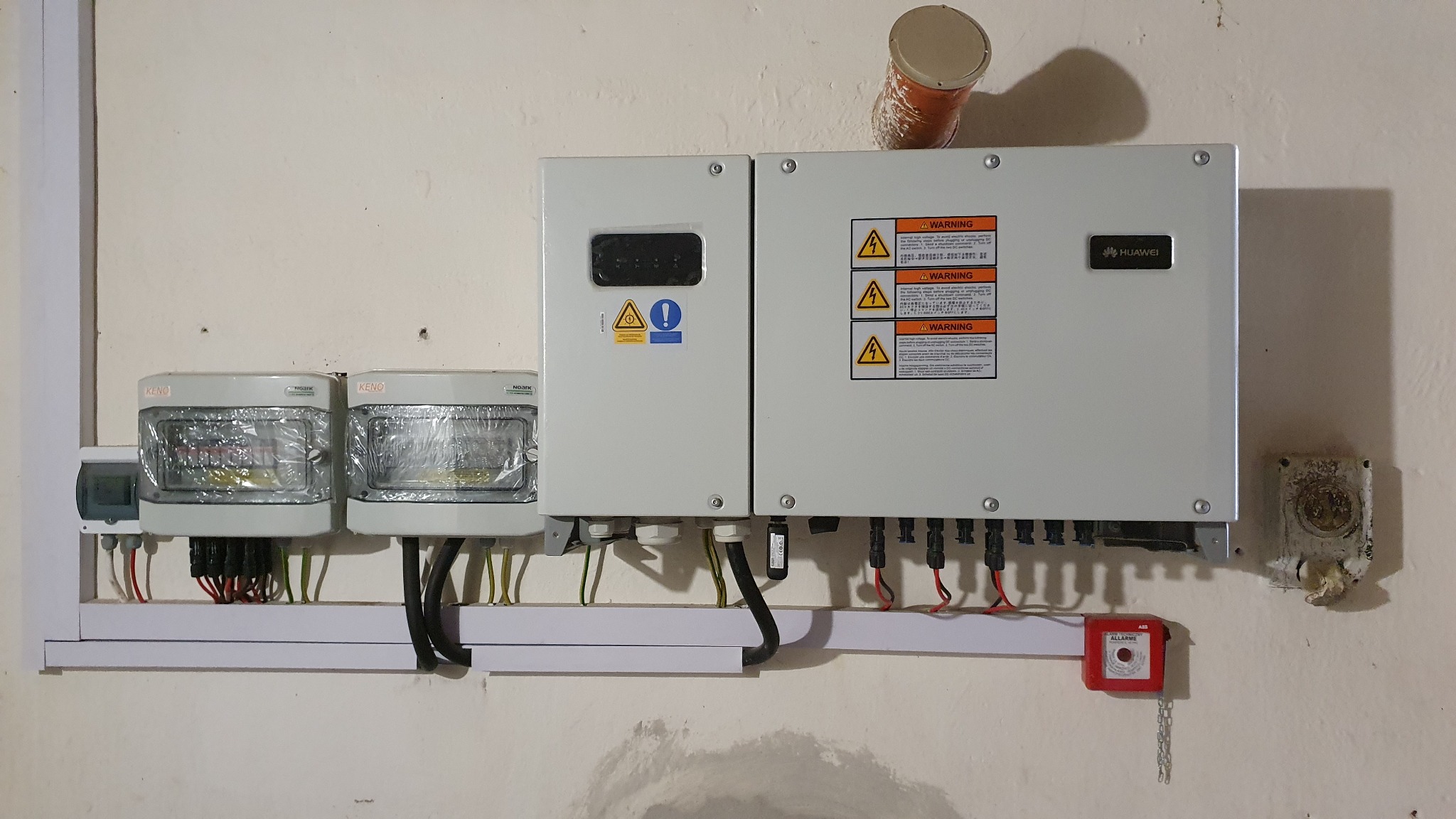 Szary falownik Huawei zamontowany na ścianie obok liczników energii elektrycznej KENO, z widocznymi przewodami, zabezpieczeniami i puszką instalacyjną.